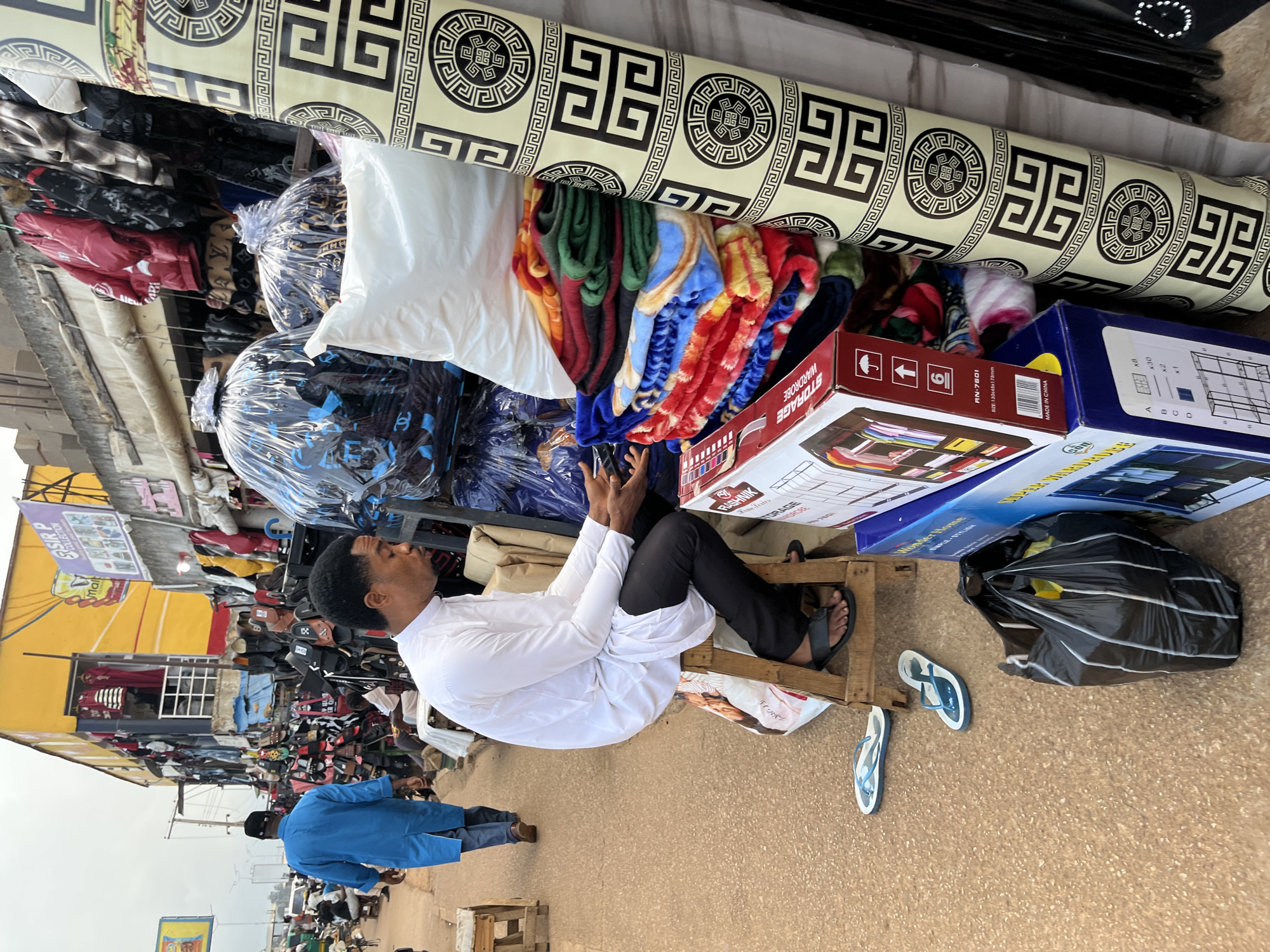 This media file captures a busy street market scene in Bukuru, Nigeria. The primary subjects are people, both actively engaged in the market and passing through. A man in a white shirt and black pants is seated on a wooden structure, possibly taking a break. Another person in a blue garment is walking away from the camera. The market is filled with various goods, including stacks of blankets, rolled-up fabric with a Greek-key pattern, packaged storage boxes, and what appear to be hats and other clothing items displayed for sale.
The setting is an outdoor market with stalls and awnings suggesting it's a commercial hub. The ground is paved, and the overall impression is one of a lively, bustling environment. The time of day is not explicitly clear but the lighting suggests it's daytime, likely overcast given the muted tones. The activity is predominantly commerce, with goods being displayed and people moving around.
Notable details include the text visible on the storage boxes, such as "RASHNIK Home Series", "WARDROBE STORAGE", "MADE IN CHINA", and model numbers like "AN-7601". There's also text on signage and the blankets, though less legible. The presence of multiple pairs of flip-flops scattered on the ground indicates casual wear and the practical nature of the environment. The scene depicts a typical day in a Nigerian market, highlighting the commerce and community interaction.