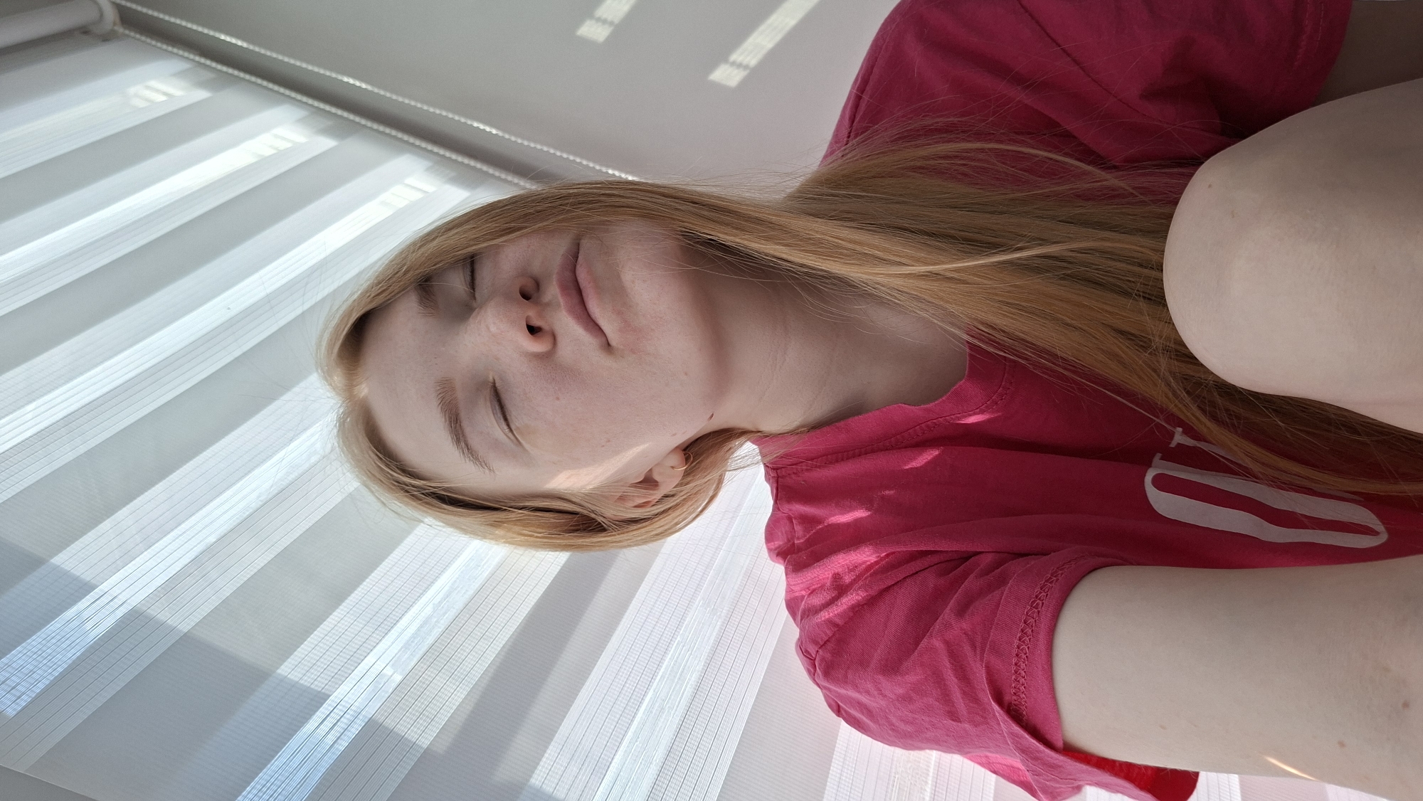 A fair-skinned, light-haired woman in a pink shirt is seen at close range from an upward angle. Her eyes are closed. The background features a white, slatted window covering. Location information for the city of Chaikovsky, Russia, is not evident in the image.
