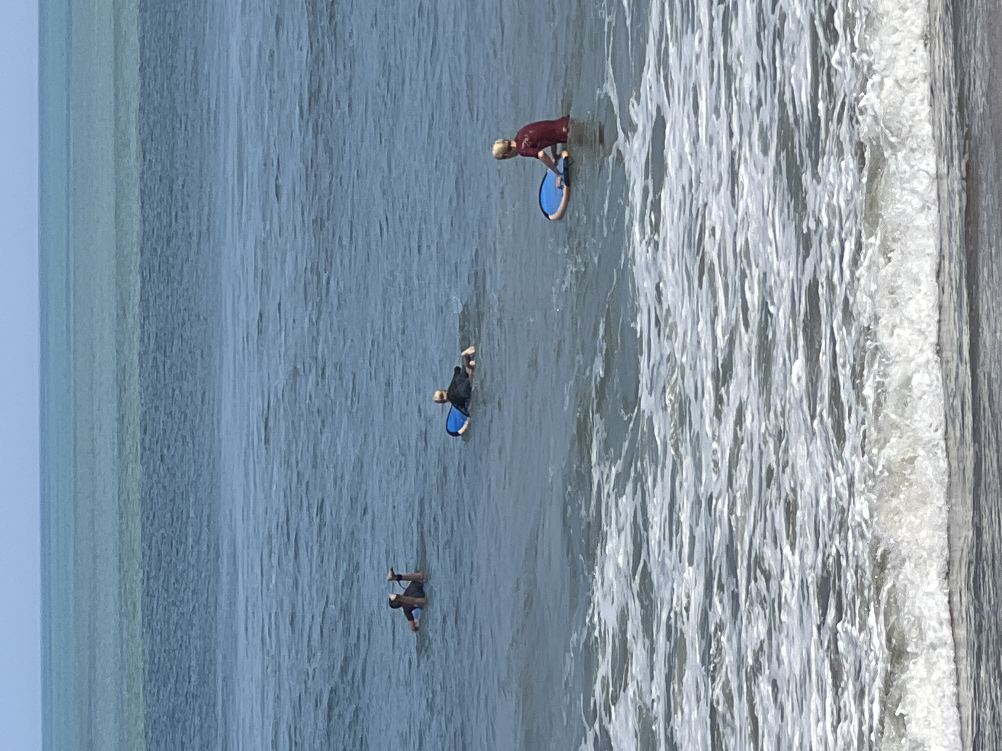 The image shows a view of the ocean off the coast of Kuta, Indonesia, on a clear day. The water is a gradient of blue and green hues, with a line of white foam from waves in the foreground. Three individuals are visible in the water, each with a boogie board. One person, wearing a maroon rash guard, is positioned in the right-center of the frame, resting on their boogie board, which is blue and yellow. To the left of this individual, further out in the water, a second person, wearing a black rash guard, is also on a blue boogie board. In the upper left quadrant, a third person is lying on their back with their legs in the air, also with a boogie board. The horizon line is visible in the distance, where the water meets a pale blue sky.