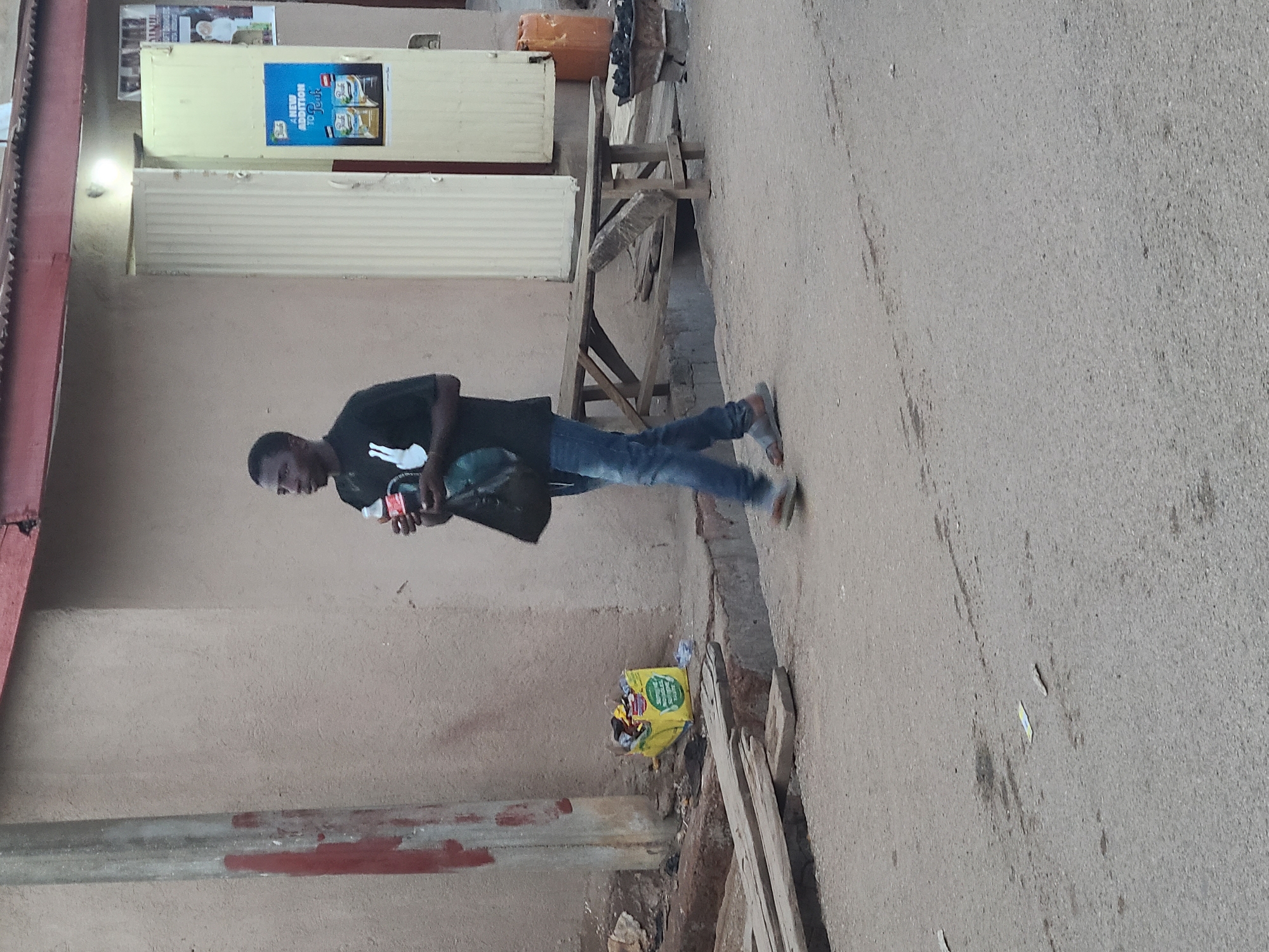 A young man walks along a light-colored, uneven paved path in what appears to be an urban or semi-urban setting, possibly a street or market area. The image is oriented vertically but depicts a horizontal scene. The man, with dark skin, is wearing a black t-shirt with a white graphic, blue jeans, and light-colored slip-on sandals. He holds a red and white container, likely a drink, in his right hand and a black bag in his left hand. His gaze is directed downwards and slightly to his left.
To the man's left is a plain light brown wall. A metal bar, partially painted red, runs along the lower section of this wall. Further along the ground, a yellow bag filled with debris or trash is visible. Behind the man, a rustic wooden bench or stall made of rough planks stands next to the wall. In the background, two light-colored, possibly cream or light yellow, roll-down security shutters or stall fronts are visible, suggesting storefronts. Above these shutters, posters are affixed to the wall. One prominent poster is blue and white, displaying the text "A NEW ADDITION TO" followed by a partially obscured word and images of two product packages. An orange jerry can and some dark objects rest on the wooden structure in the midground. The lighting suggests daytime, possibly overcast, providing soft, natural illumination. No specific weather conditions are discernible, and the man's expression is neutral.