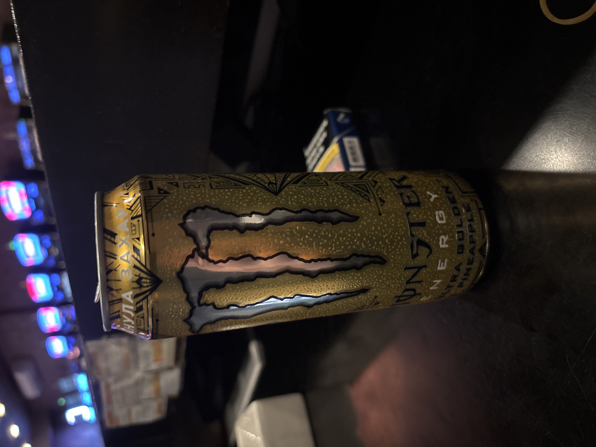 A horizontal image depicts a yellow and gold Monster Energy Ultra Golden Pineapple can lying on a dark surface. The can has the recognizable monster claw logo on its front. In the background, there is a stack of white boxes and blue and white packaging, possibly cigarettes, stacked and organized in a disorderly fashion. Beyond the immediate area, the image captures a colorful blur of lights, potentially from electronic devices. It is not possible to confirm the location as Plovdiv, Bulgaria.