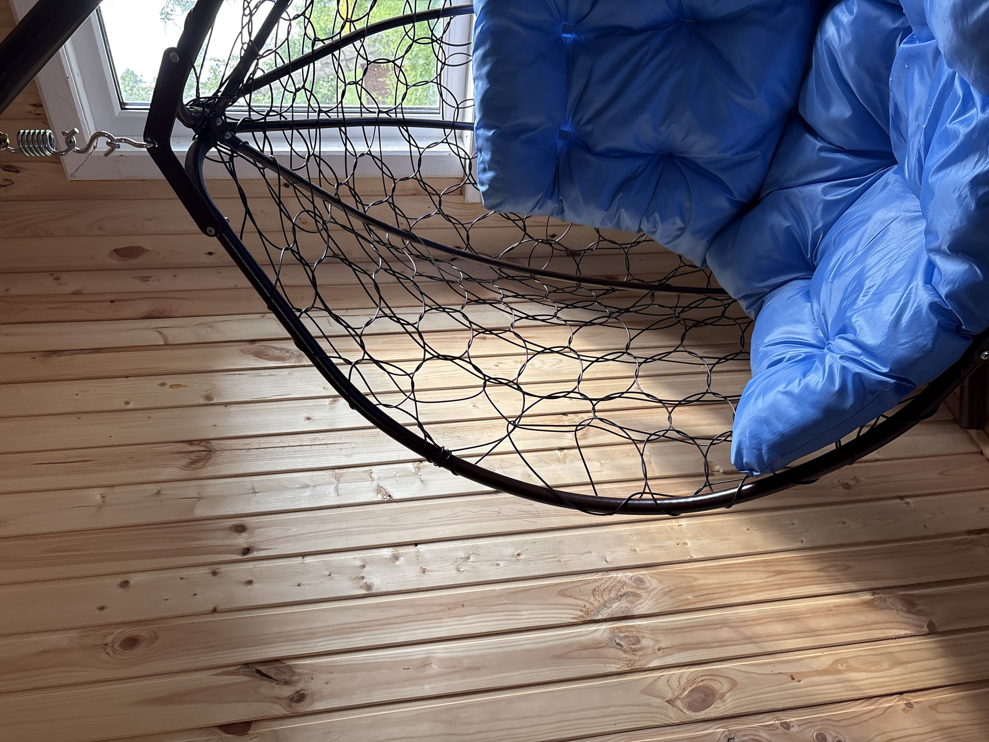 The image shows a close-up of a blue cushioned hanging chair in a room with light-colored wooden paneled walls. The chair is suspended by a metal spring and hook, visible at the top of the frame. The chair's frame is dark metal with a woven pattern. A window with green foliage visible outside is partially seen to the right of the chair. The scene is indoors and appears to be a domestic setting, possibly in a residence in Italmas, Russia, though there is no direct evidence to confirm the location.