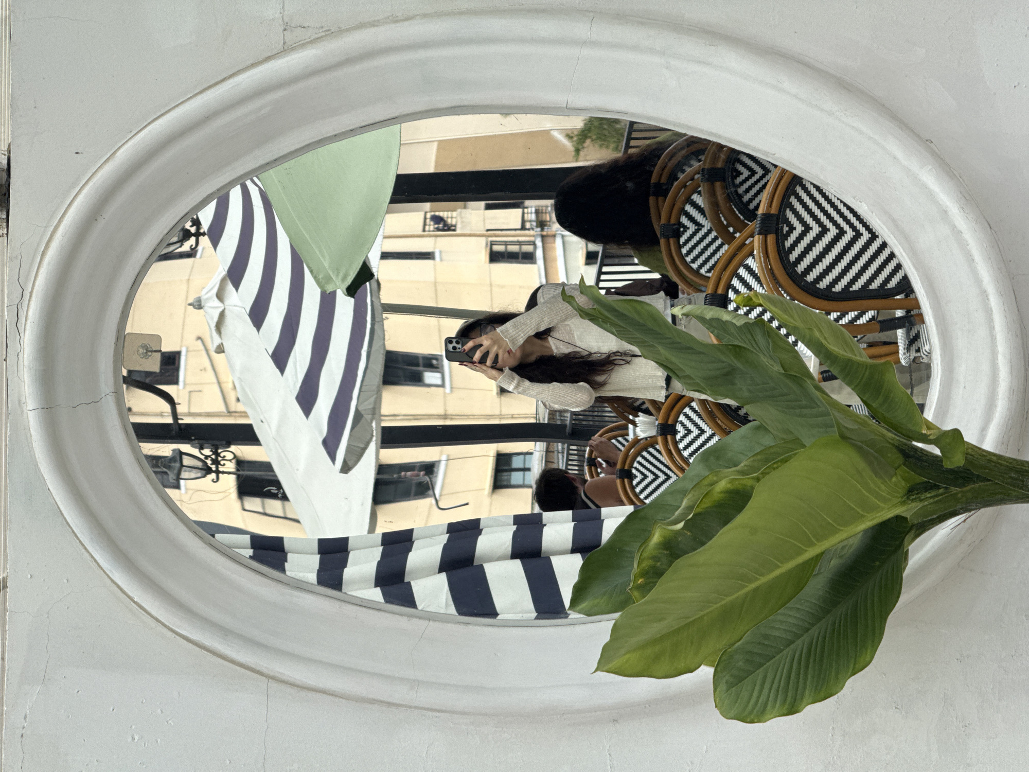 The image is a reflection in an oval mirror, set into a white stucco wall. The mirror shows an outdoor seating area. In the foreground of the reflection, a woman is taking a selfie with her phone. She has long dark hair and is wearing a light-colored, textured sweater. She is seated at a table with black and white chevron patterned chairs. Behind her, another person with dark hair is seated at a table. Several umbrellas are visible in the background, one with navy and white stripes and another light green one. Buildings with yellow facades and dark windows are visible behind the umbrellas. A street lamp with an ornate design is also reflected in the mirror. A large green plant with broad leaves is in the foreground of the actual image, partially obscuring the mirror's left side. The scene appears to be a cafe or restaurant with outdoor seating in Da Lat, Vietnam, suggested by the style of architecture and outdoor dining common in the city.