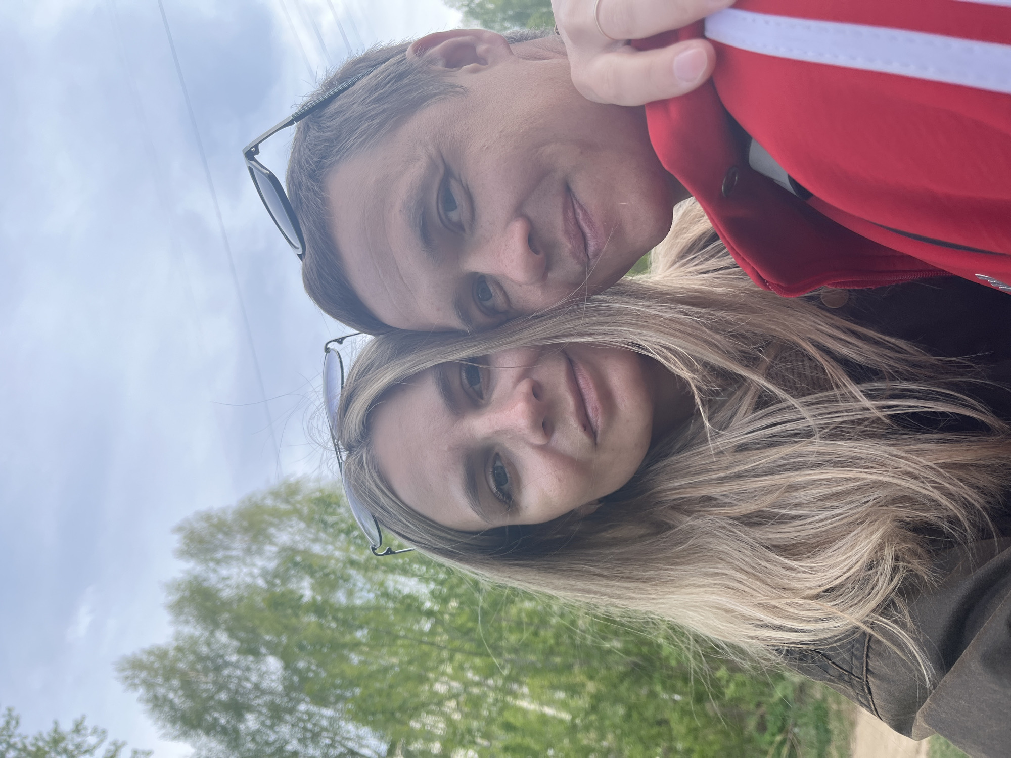 A close-up selfie shows two individuals, a male and a female. The male is wearing a red jacket with white stripes, while the female has long blonde hair and sunglasses perched on her head. Both individuals are looking directly at the camera with a slight smile. The background consists of green trees and a cloudy sky. The location is the city of Chaikovsky, Russia.
