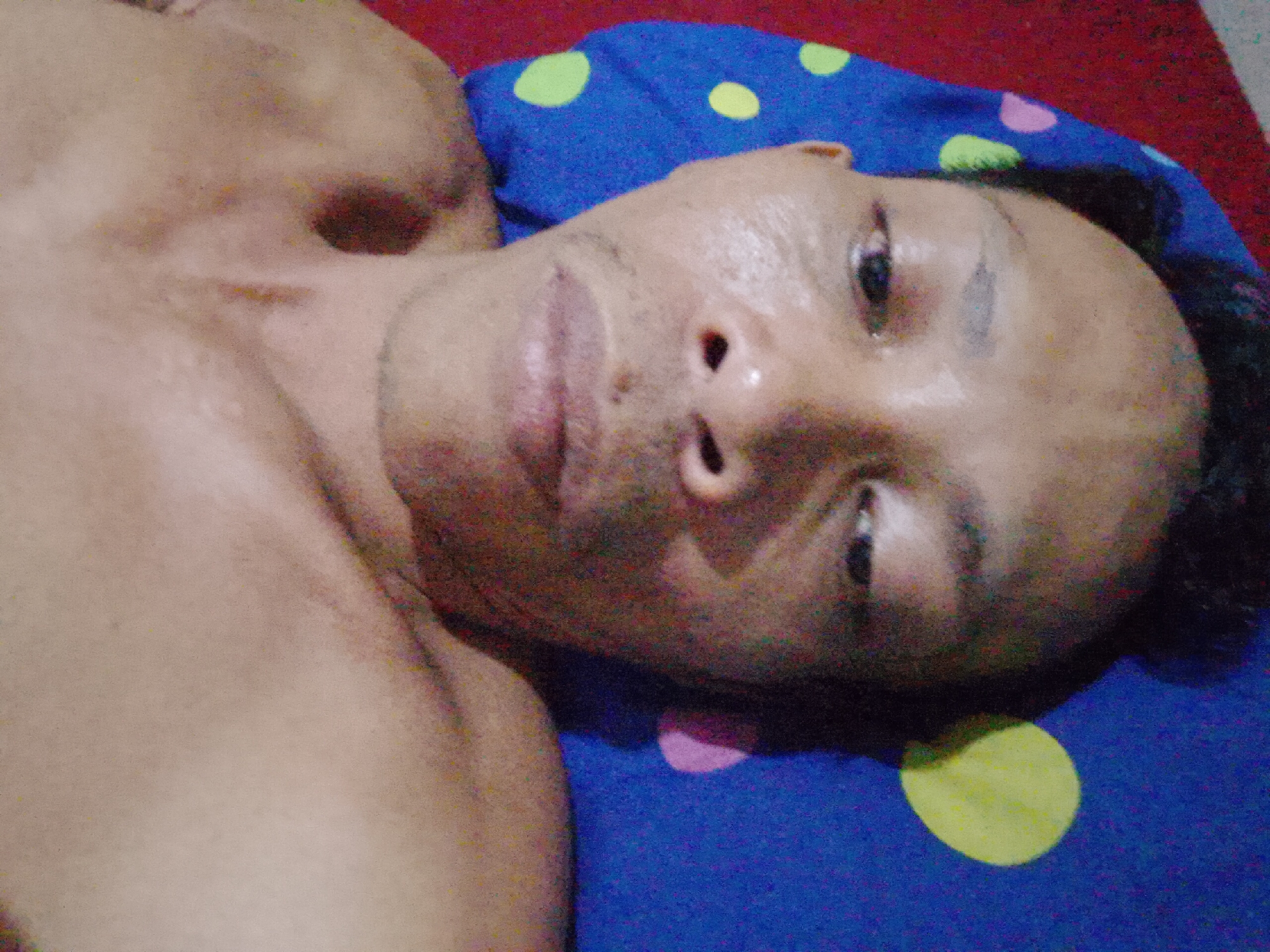 A shirtless man is lying on a blue polka-dot pillow on what appears to be a red surface. The man is light brown skinned with dark hair. The location is Bukit Ketok, Indonesia.