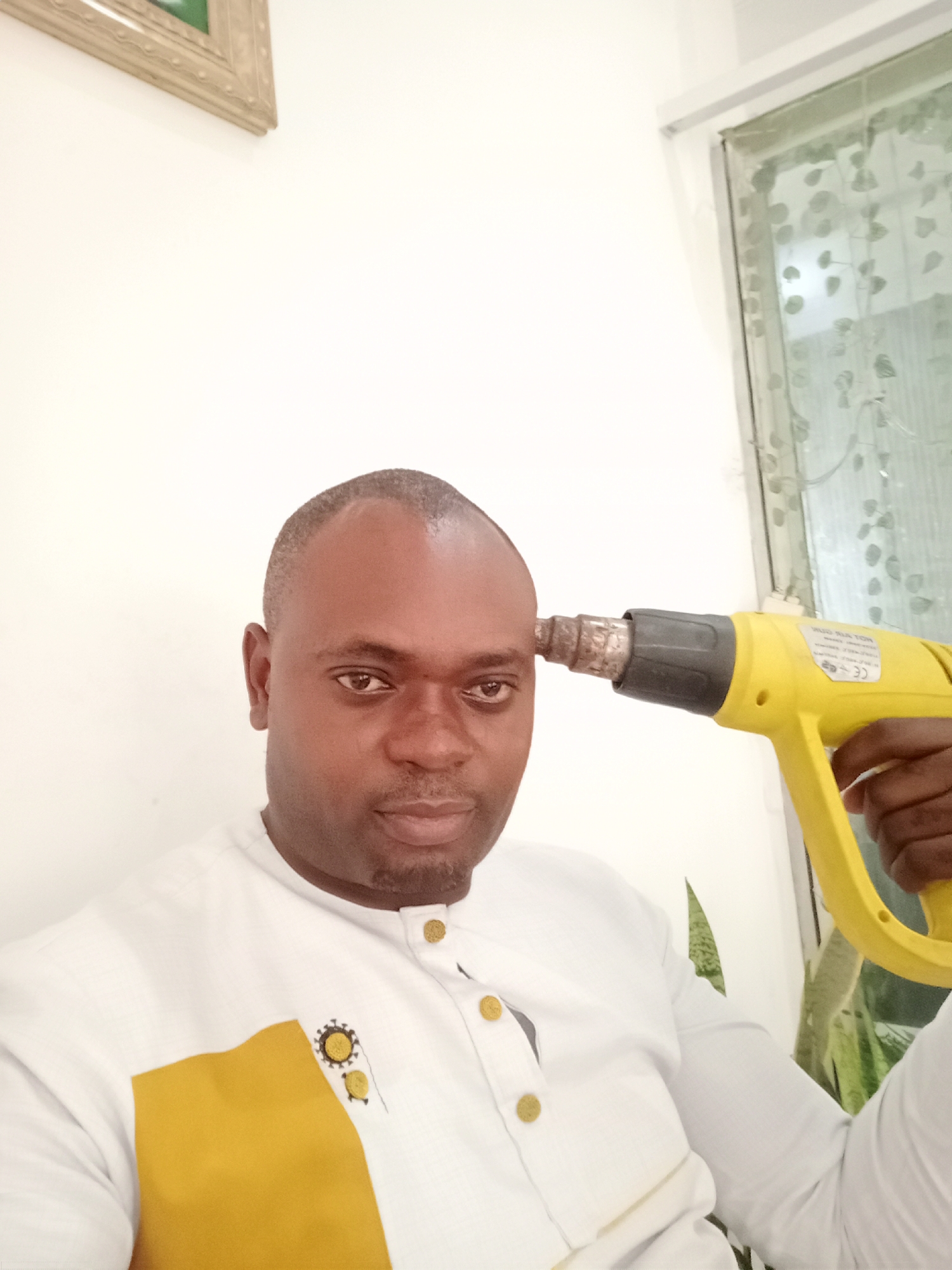 The image captures a dark-skinned man indoors in Gwarinpa, Nigeria, looking directly at the camera with a neutral expression. He has a shaved head and a short beard. He is wearing a long-sleeved white shirt with a mustard yellow patch on the left chest, accented with small gold buttons and black embroidered designs resembling suns or gears.
The man is holding a yellow and black heat gun to the left side of his head, near his temple, with the metallic nozzle touching his skin. Visible text on the heat gun includes "SUON" (possibly part of a brand name) and a "CE" mark.
The background features a plain white wall, with a light-colored framed picture partially visible in the upper left corner. To the right, there's a window with frosted or patterned glass adorned with a leaf-like design, through which some green foliage can be seen. The lighting in the scene is bright and even.