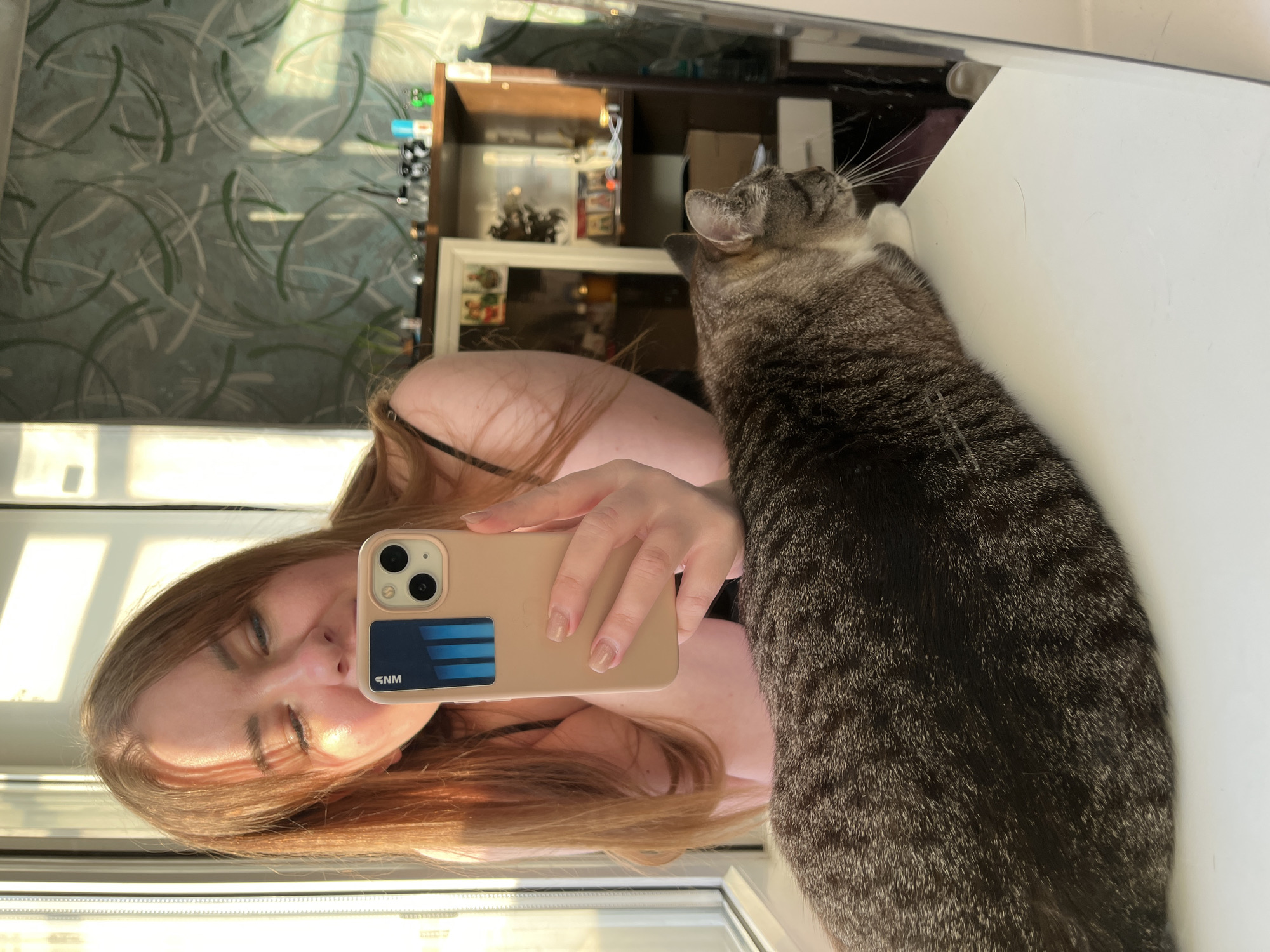 A mirror selfie of a woman with long auburn hair and a cat. The woman is holding a beige smartphone with a blue and black card attached to the back. The cat is a tabby with gray and black fur. It is sitting on a white surface. The background includes a window, a wall with patterned wallpaper, and a shelf with various items. The image was taken indoors. The location is in the city of Chaikovsky, Russia.