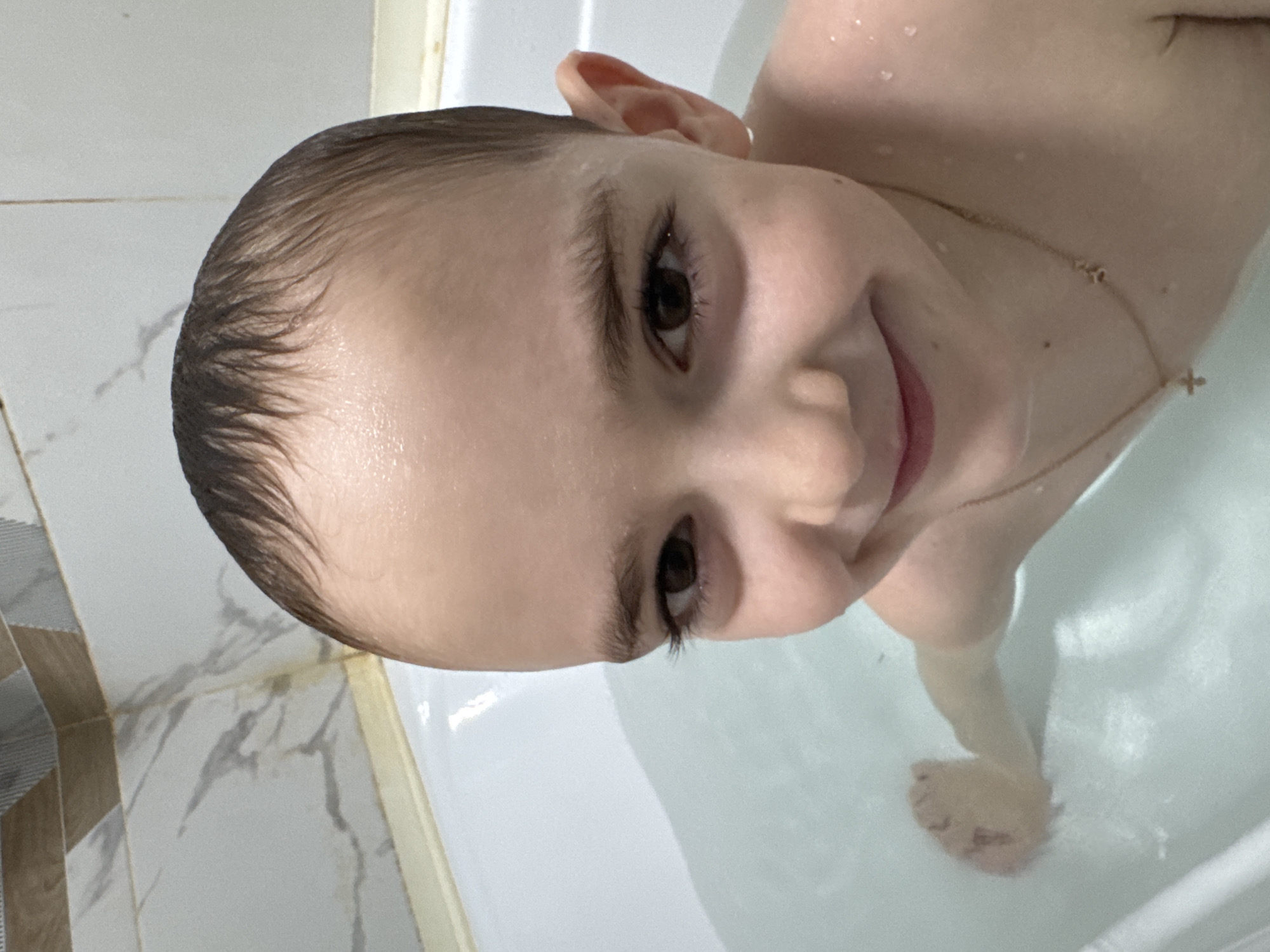 A close-up portrait captures a young child in a bathtub, smiling gently at the viewer. The child, with wet, dark hair and water droplets on their skin, is partially submerged in water, suggesting they are taking a bath. Their dark eyes and eyelashes are prominent, and a thin golden chain with a small cross pendant hangs around their neck.
The setting is a bathroom, with white tiled walls in the background. Some of the tiles feature a subtle grey marbled pattern, while another section shows beige and white rectangular tiles. The white bathtub is filled with clear water, and the child's arm, leg, and foot are visible beneath the surface. The child appears happy and content. There is no visible text in the image. The location context indicates this scene is in Раздолье, Russia.