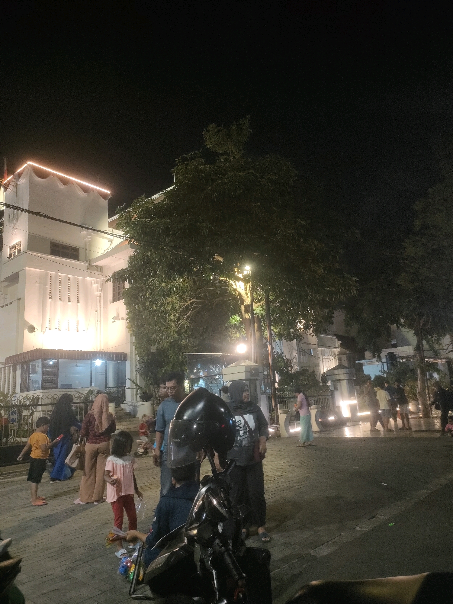 The image depicts a night scene in Surabaya, Indonesia, likely in a public area with a building and several people. In the foreground, a motorcycle is partially visible. A young girl in a pink shirt and red pants is standing in the middle of the paved area, holding some items. To her left, a man wearing a blue shirt stands beside her, and a woman in a maroon hijab and brown pants is seen slightly behind them. Further to the left, a young boy in an orange shirt and black shorts stands holding a toy.
The background features a large, multi-story building with white walls illuminated by warm-toned lights. The building has a distinctive architectural style with patterned facades and decorative lighting along its upper edge. A large tree with dense foliage stands to the right of the building, partially obscuring it. Several other people are scattered throughout the scene, walking or standing. The ground is paved with interlocking bricks. The overall atmosphere suggests a lively evening gathering or a marketplace.