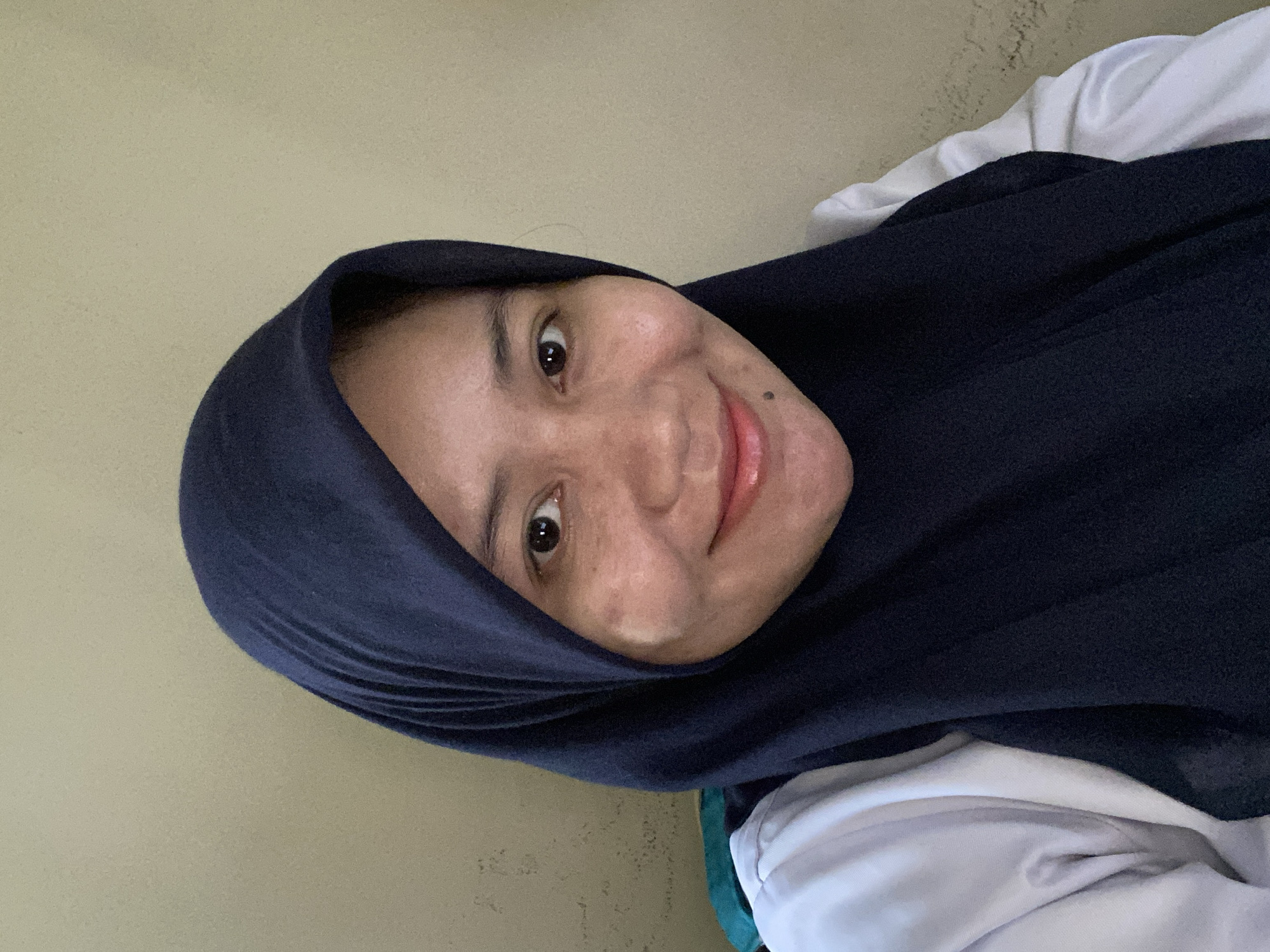 A headshot image of a young Indonesian woman wearing a dark blue hijab and a white collared shirt is presented. The woman is positioned in the center of the frame, facing the camera directly, and is smiling. Her left cheek is slightly obscured by the hijab. The background is a plain, textured wall in a light tan color. The lighting is soft, casting a warm glow on the subject's face. The image appears to have been taken indoors, possibly in a residential or office setting in Banjarbaru, Indonesia. There are no other people or objects visible in the frame.