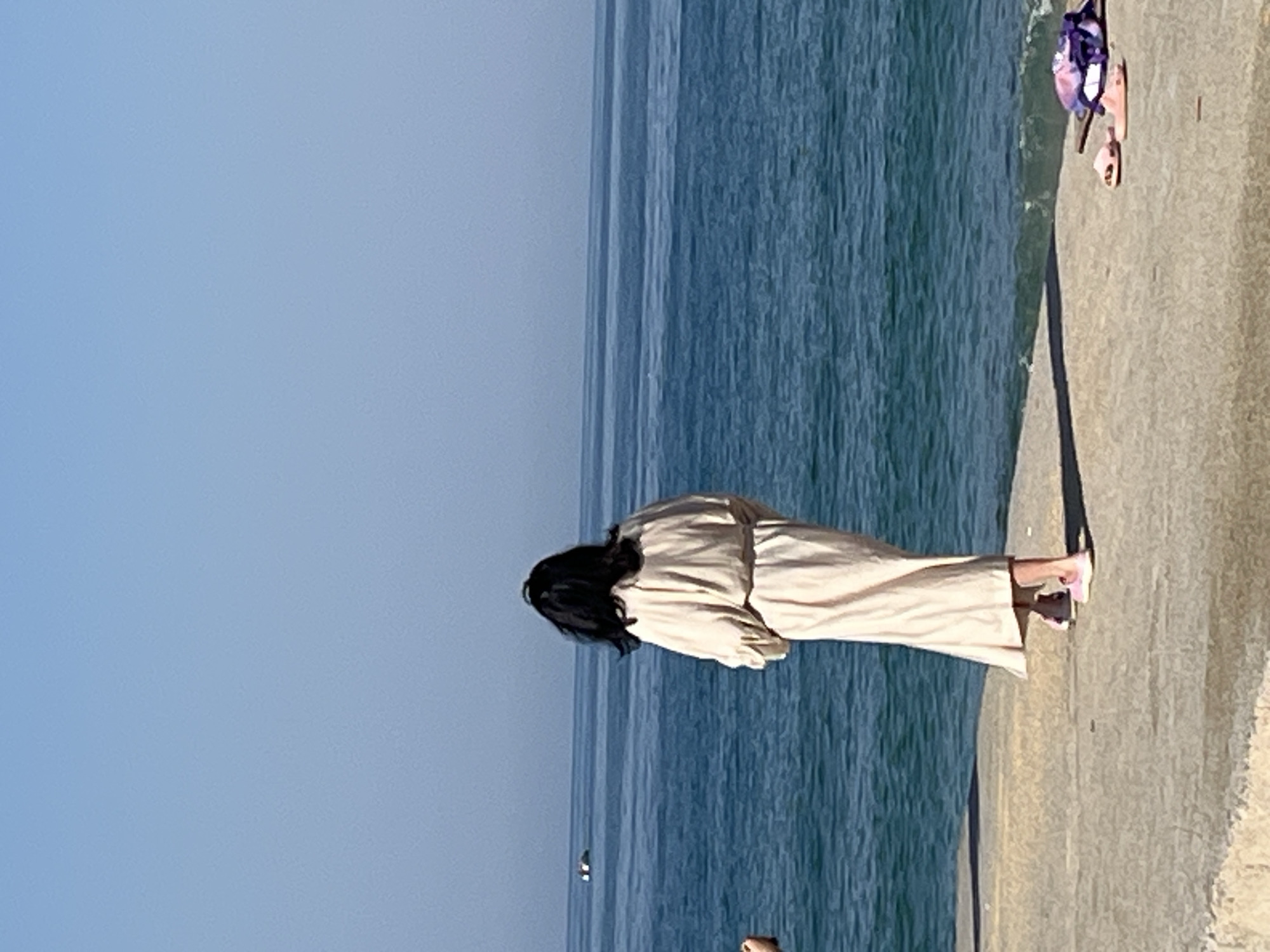 A woman stands on a concrete walkway beside the sea. She has long, dark hair, and is wearing a light-colored dress or robe. The sea is a deep blue color, and the sky is a lighter blue. The walkway is a light gray color. In the background, there is a small island in the sea. On the edge of the walkway, a bag and a pair of sandals can be seen. The location appears to be Alanya, Turkey, given the coastal landscape and the presence of the island in the distance.