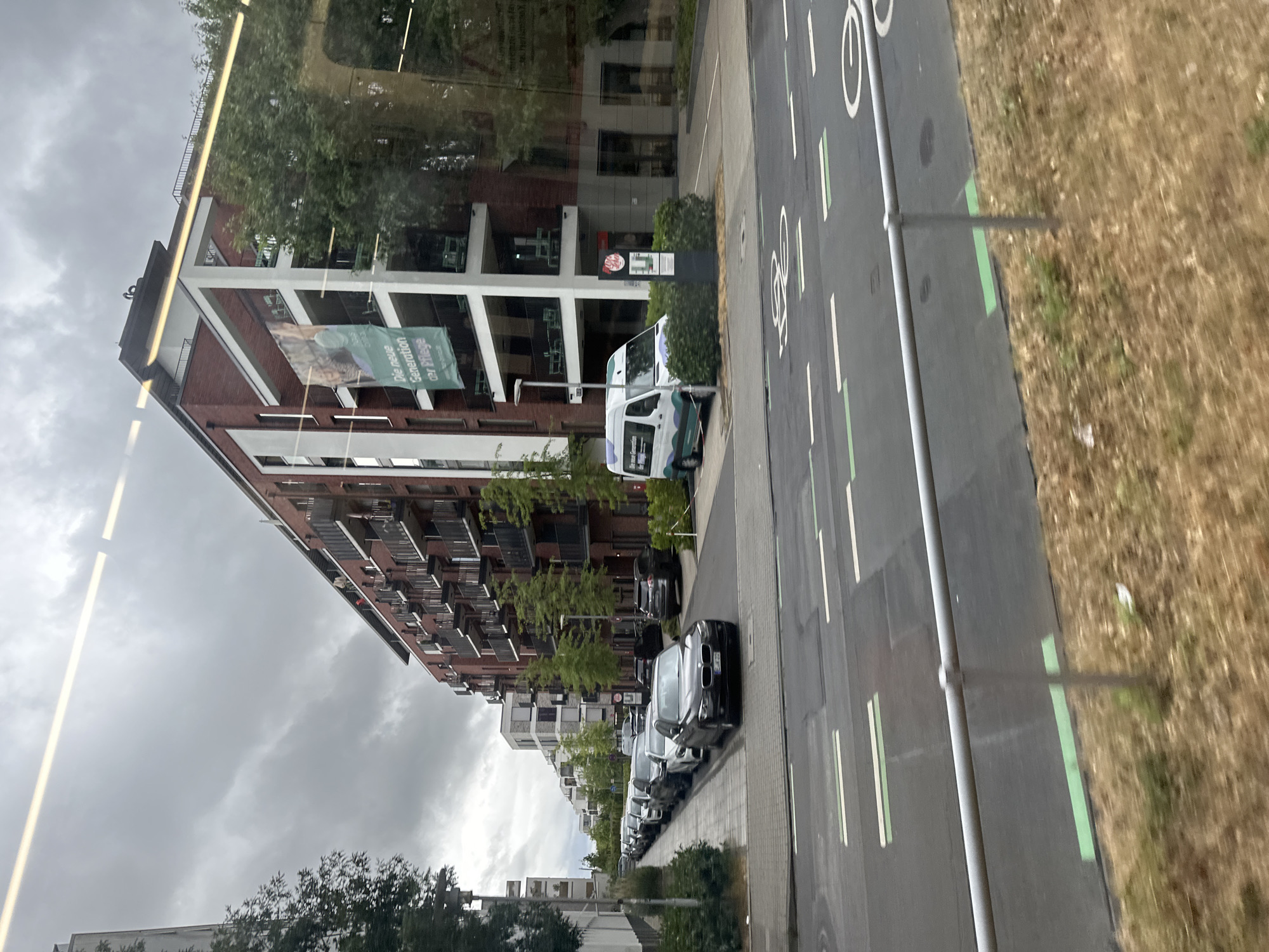 This image depicts a street scene in Dusseldorf, Germany. On the right side of the image, a modern brick apartment building with balconies and large windows dominates the frame. A banner with German text "Die neue Generation der Pflege" and images of people is visible on the building. In front of the building, a white van with a green and purple design is parked. A street lamp and a signpost are also present. The street in the foreground has markings for a bike lane, including bicycle symbols and green lines. Several cars are parked along the sidewalk to the left of the street, and more apartment buildings are visible further down the street. The sky is overcast and cloudy, and the ground is covered with dry grass.