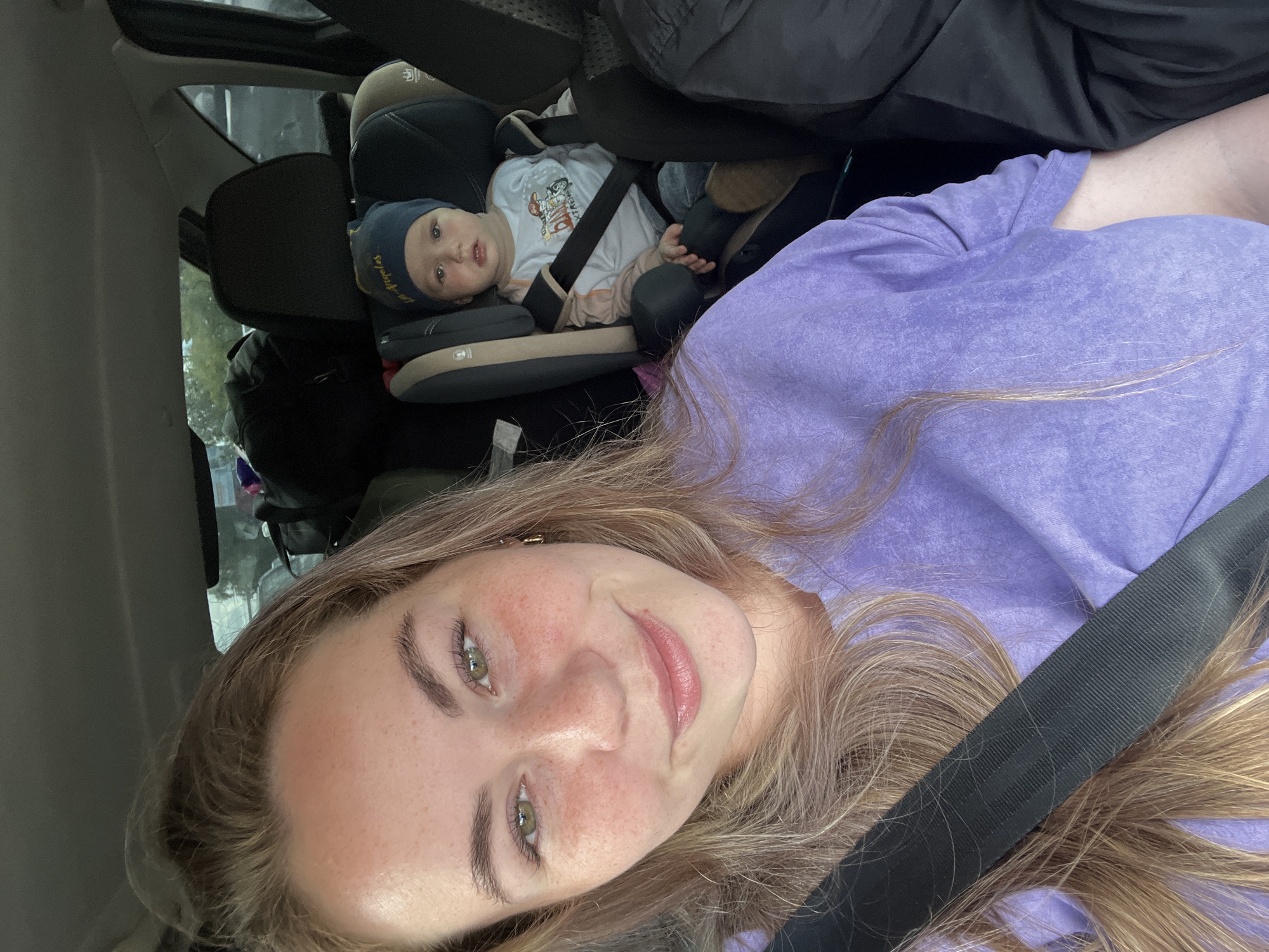 A woman with blonde hair and freckles is taking a selfie in a car. She is wearing a purple sweatshirt and a seatbelt. Next to her, in a car seat, is a baby wearing a blue beanie with "Los Angeles" written on it and a white shirt with graphics. The baby is looking at the camera. The car appears to be packed with luggage in the background.