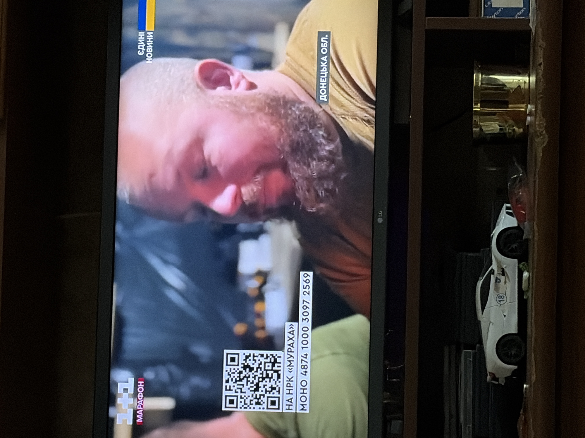 The image shows a television screen displaying a news broadcast. The broadcast features a man with a beard and a crew cut, wearing a yellow t-shirt. Text overlays on the screen include "1+1 МАРАФОН" in the upper left corner, "ЄДИНІ НОВИНИ" with a Ukrainian flag to the right, and "ДОНЕЦЬКА ОБЛ." in the lower right. A QR code is also visible in the middle left, with the text "НА HPK «МУРАХА»" and a phone number "МОНО 4874 1000 3097 2569" below it. The television itself is placed on a shelf, with a white toy car and a gold cylindrical object visible on the shelf in front of it. The background appears to be a dimly lit room.