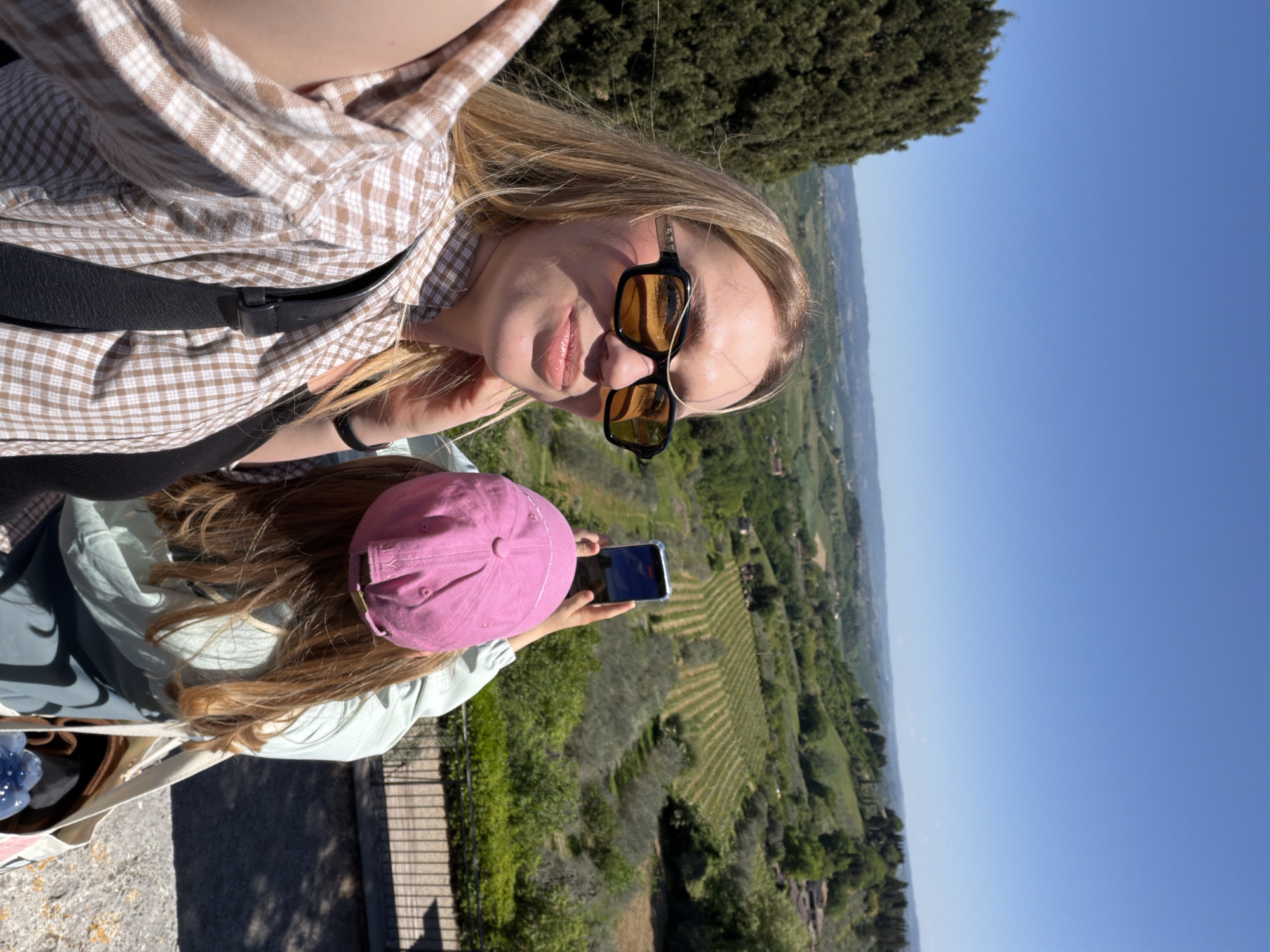 The image shows two young women in San Gimignano, Italy, overlooking a green valley under a clear blue sky. The foreground woman, who is taking the selfie, is wearing a brown and white checkered shirt, a black shoulder strap, and black sunglasses. The second woman, standing behind her, is wearing a pink baseball cap and a light green jacket, and she's holding a phone to take a picture of the valley below. The location seems to be an elevated point within or near San Gimignano, providing a wide view of the Tuscan landscape.