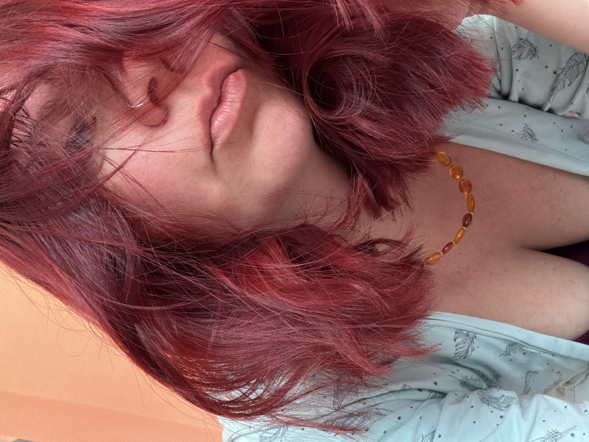 A close-up, low-angle portrait features an individual, presumably female, situated indoors in Chaikovsky, Russia. The subject's face is partially obscured by vibrant red-magenta hair that cascades over the forehead, nose, and cheeks. A small, silver hoop nose ring is visible in the subject's right nostril. The lips are closed, exhibiting a natural light pink hue. The subject wears a light blue garment, possibly a robe or pajama top, patterned with dark feathers and small dots. This garment is open, revealing the upper chest and cleavage, where a necklace of irregularly shaped, polished amber and reddish-brown beads is positioned. Freckles are visible on the skin of the upper chest. A dark garment is partially visible beneath the light blue one. The background is a plain, uniform light orange-peach colored wall.