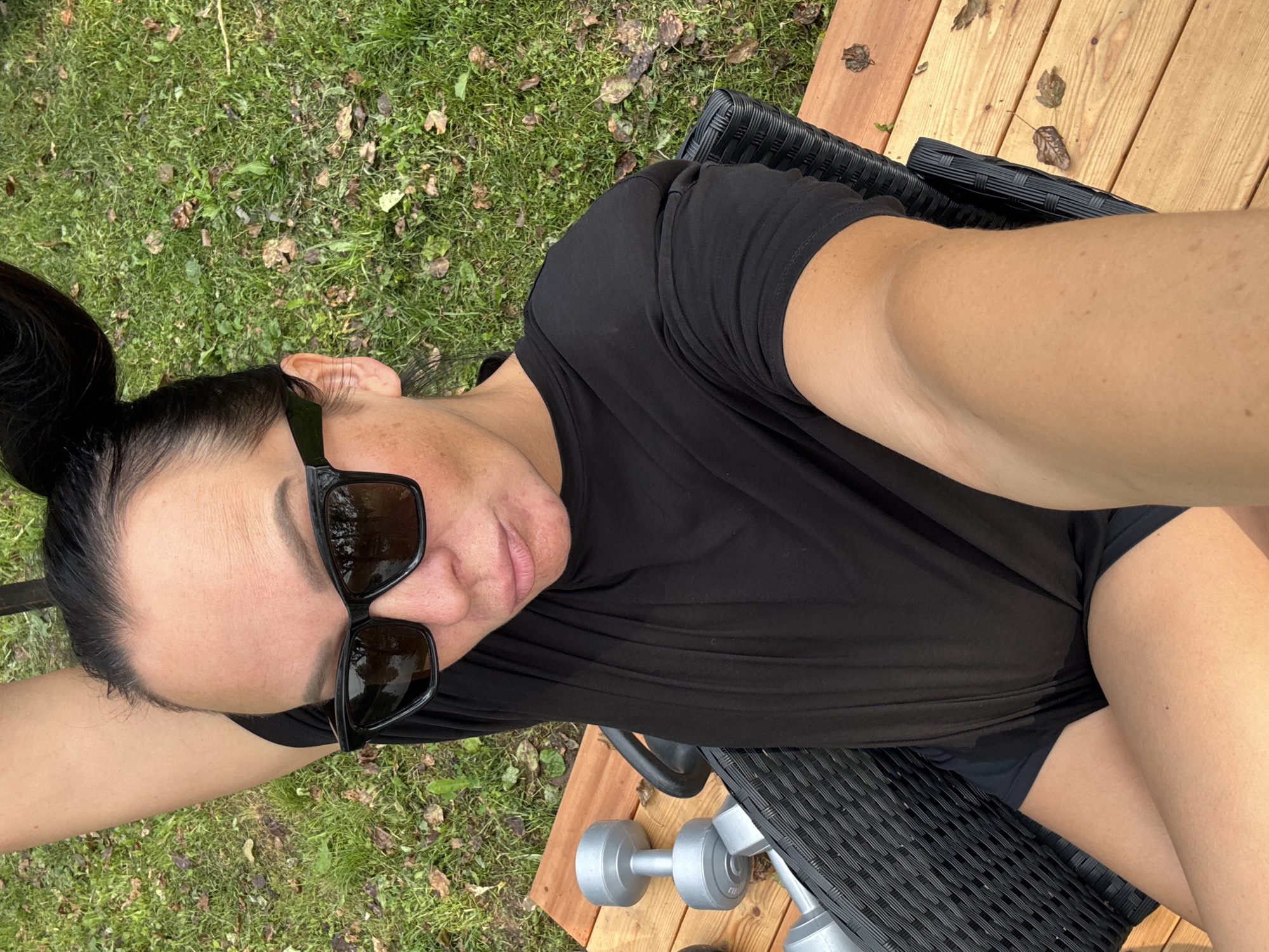 A person with dark hair pulled into buns is sitting on a wicker chair on a wooden deck. They are wearing sunglasses and a black t-shirt with black shorts. Two silver dumbbells are visible on the deck. The background is a grassy area with some fallen leaves. The image was taken outdoors, likely in a residential area. The city of Chaikovsky, Russia is not visible in this image.