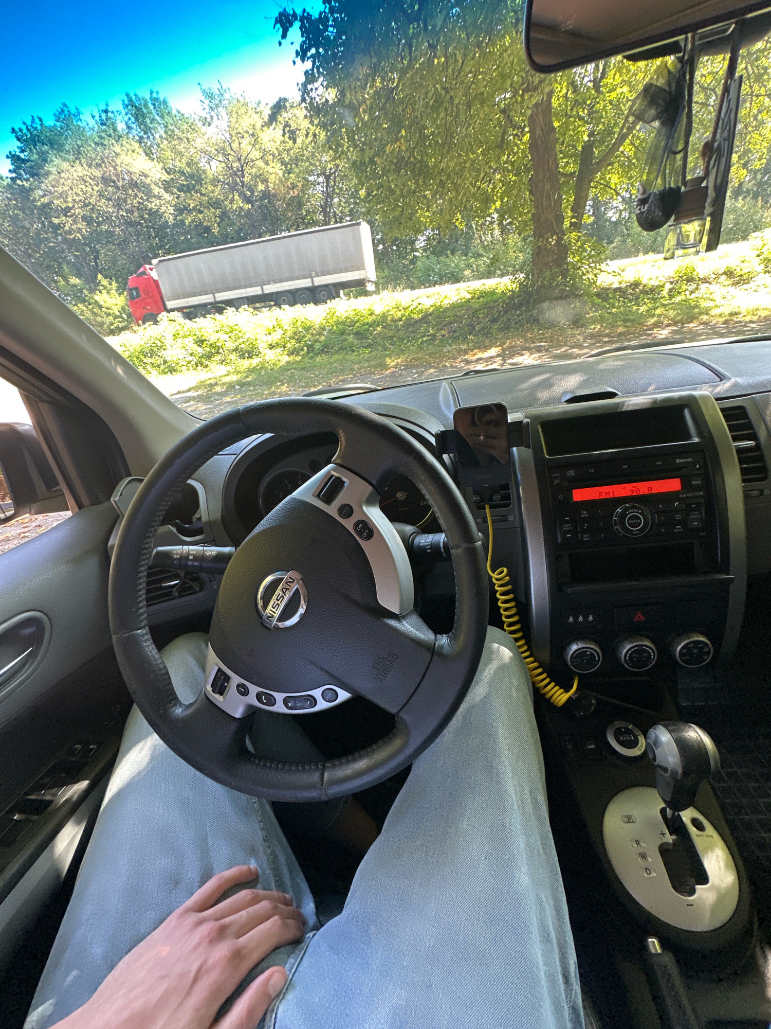 The image is taken from the perspective of someone sitting in the driver's seat of a car. The steering wheel is prominently in the foreground, with the Nissan logo visible. To the right of the steering wheel is the car's dashboard, featuring a radio display showing "FM1 90.0" and various buttons and knobs. A yellow coiled cable is attached to a phone mount on the dashboard. The driver's legs, clad in light blue jeans, are visible. Through the car's windshield, a bright, sunny outdoor scene is visible. In the background, a red truck cab is attached to a long, silver trailer, parked or stopped on the side of a road lined with green trees and foliage. The setting appears to be a rural or semi-rural area with lush vegetation.