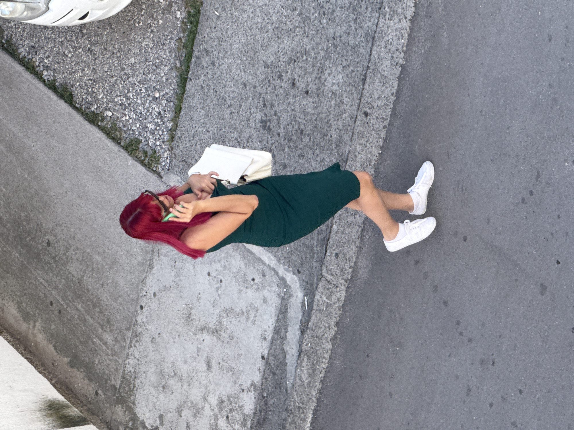 An overhead, medium shot captures a woman with vibrant red hair, wearing a dark green dress and white sneakers, standing on an asphalt road in Budva, Montenegro. She is holding a cell phone to her left ear and a white purse in her right arm. Her sunglasses are perched on her nose. To the upper left of the frame is a concrete walkway, and to the upper right is a gravel-covered area adjacent to a white vehicle. The scene appears to be outdoors during daylight.