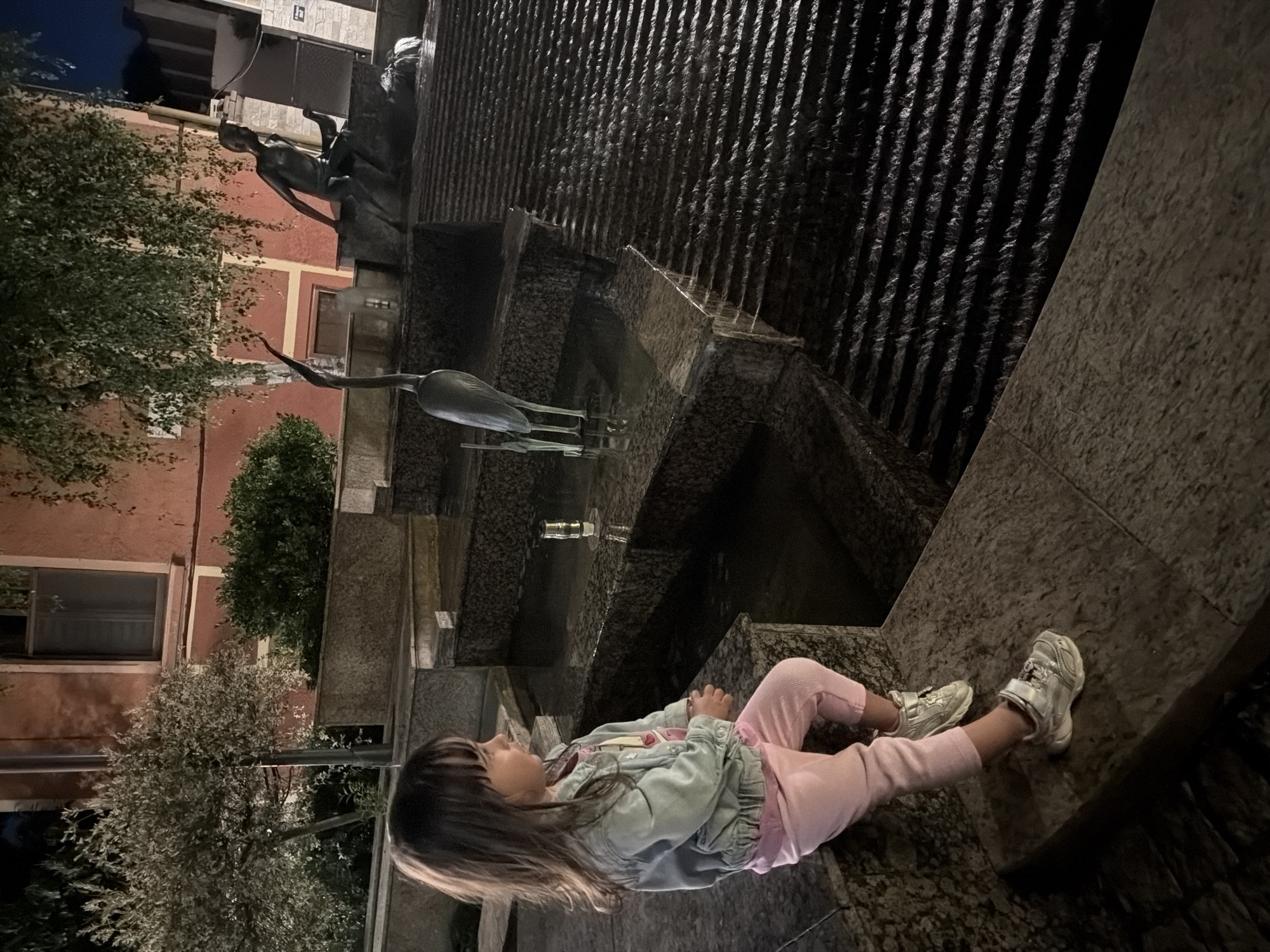 A young girl sits on the edge of a multi-tiered stone fountain, which has water flowing down its ribbed surface. The girl is wearing a light blue jacket over a pink shirt, and pink pants. She has long, dark hair and is looking to her left. She is wearing silver sneakers. The fountain appears to be made of dark stone with a rough texture. In the background, there are buildings with orange and white facades, and lush green trees. A bronze sculpture of a person is visible to the left, and what appears to be a bronze heron sculpture is in the water of the fountain. The scene is dimly lit, suggesting it is either dusk or nighttime. The location is Burgas, Bulgaria.