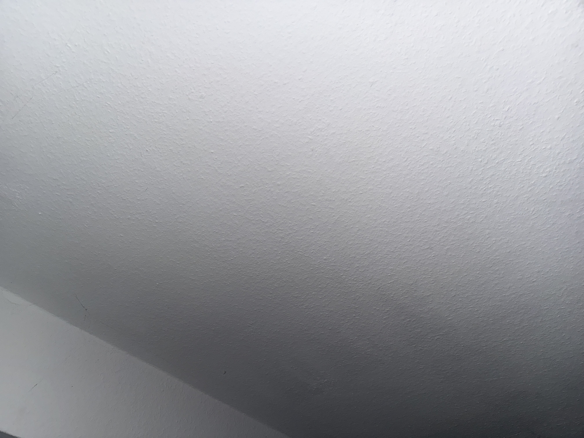 The image shows a close-up of a textured, light gray wall. The wall is illuminated from the right, creating a gradient of light and shadow that runs diagonally from the top left to the bottom right. The texture of the wall appears to be slightly rough or stucco-like. There are a few faint, thin, dark lines visible on the surface of the wall, which could be scratches or cracks. The lighting suggests the presence of a light source outside the frame to the right, casting a shadow and creating a subtle sense of depth. The overall impression is one of an indoor wall surface, possibly in a dimly lit corner or hallway due to the shadow. There are no people or objects clearly discernible in the image.