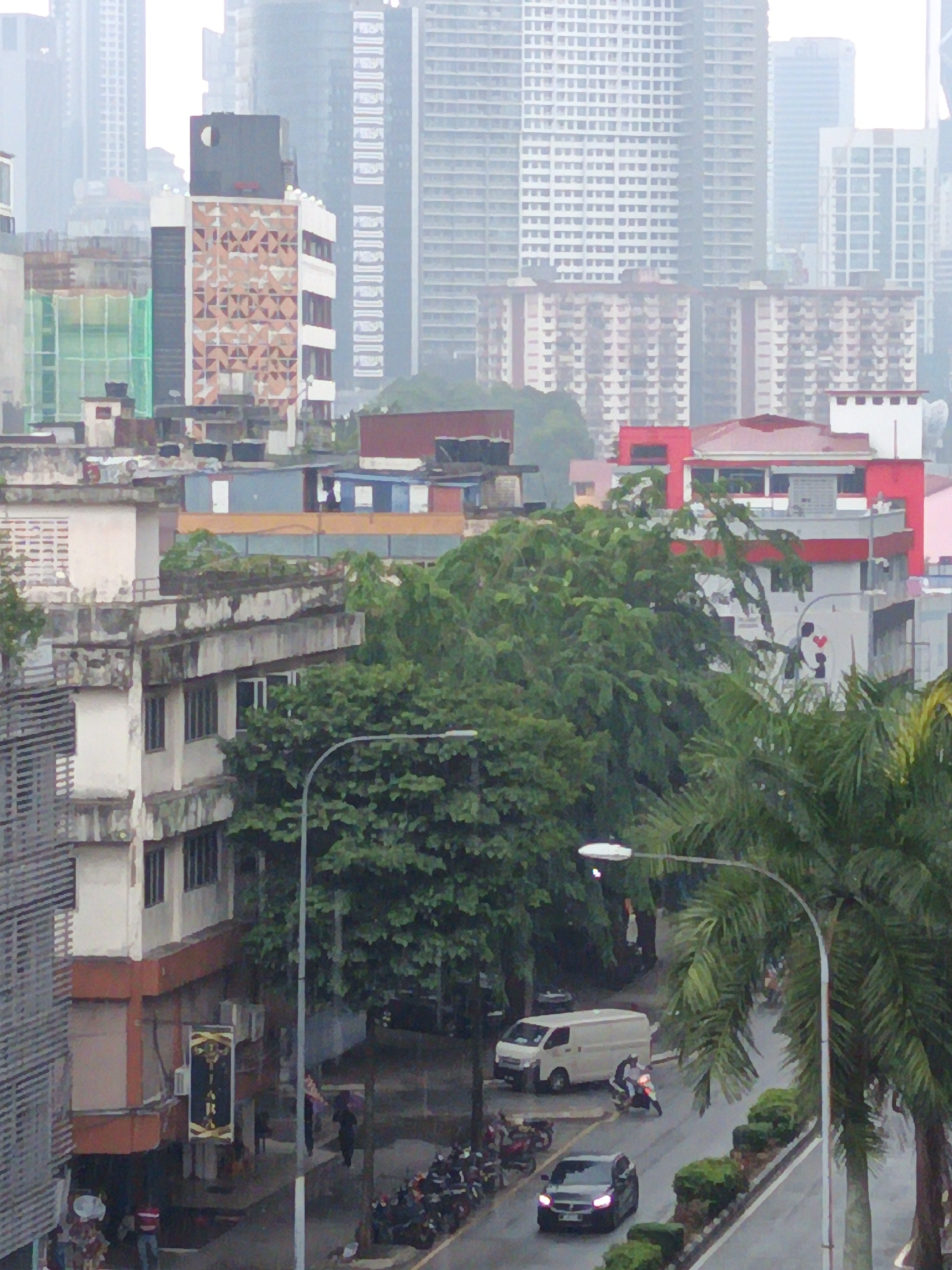This is a high-angle, full shot of a street scene in Kuala Lumpur, Malaysia, under overcast skies. The foreground features a multi-story building with shops on the ground floor, one displaying a vertical sign with gold lettering on a black background. To the right, a street curves into the distance, lined with trees and lush foliage. A street lamp arches over the road. On the street, a white van is parked, and a motorcyclist is riding past. Several motorcycles are parked along the side of the road further down. A dark sedan is also visible on the road. In the background, numerous tall buildings, some appearing to be under construction with green netting, dominate the skyline, indicative of a dense urban environment. A few individuals are present on the sidewalks, one holding an umbrella. The overall atmosphere is subdued, possibly due to the weather.