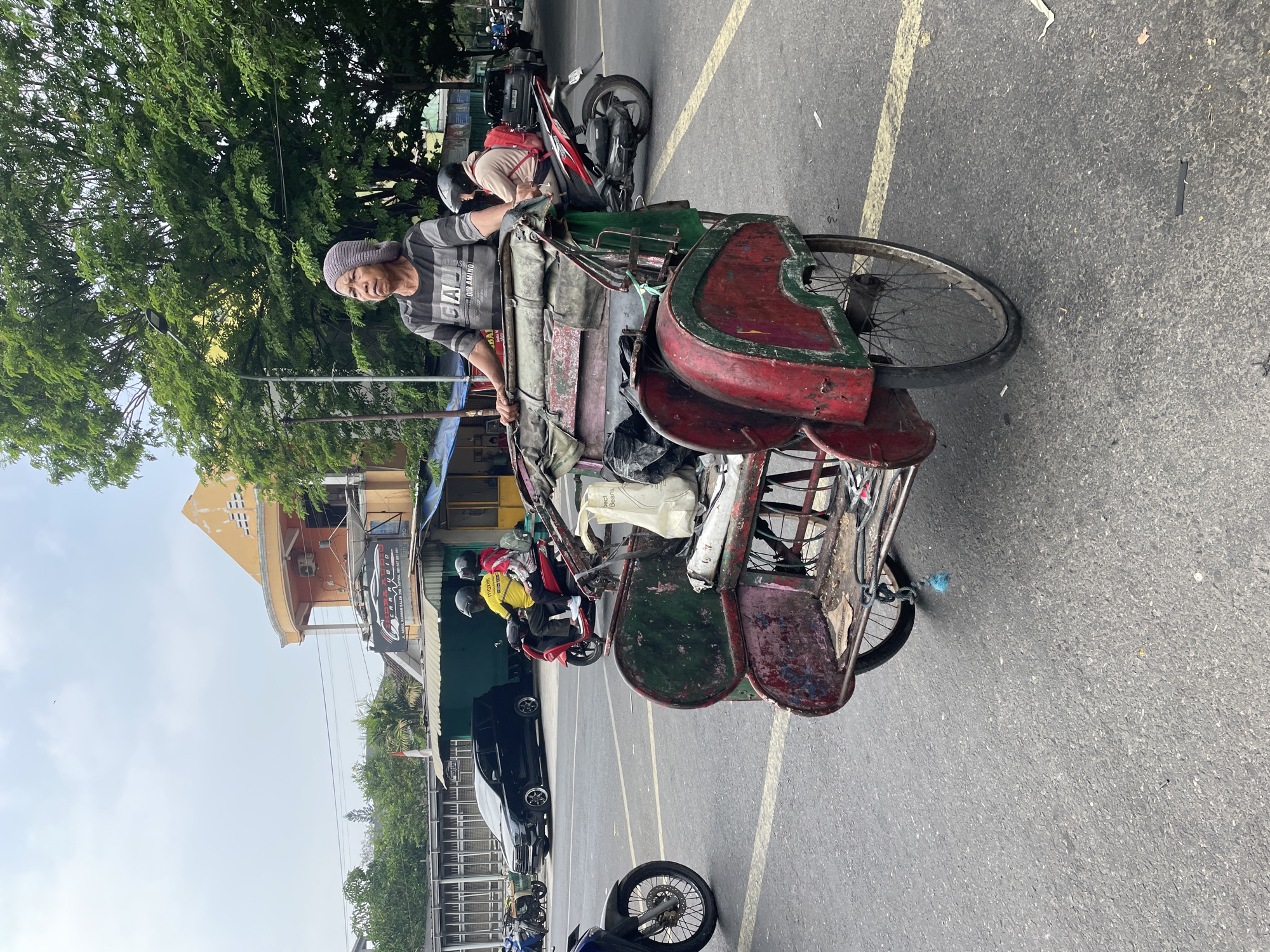 An elderly person wearing a gray knit hat and a horizontally striped t-shirt stands next to a weathered, red and green pedal-powered rickshaw on an asphalt road. The rickshaw has a worn canvas covering and appears to be carrying various items. In the background, a person wearing a yellow shirt and a helmet rides a red scooter, followed by another person in a light-colored jacket with a red backpack. Further back, a black car is parked. Buildings with orange and green facades are visible, along with lush green trees and a cloudy sky. The scene is a street in an urban environment.