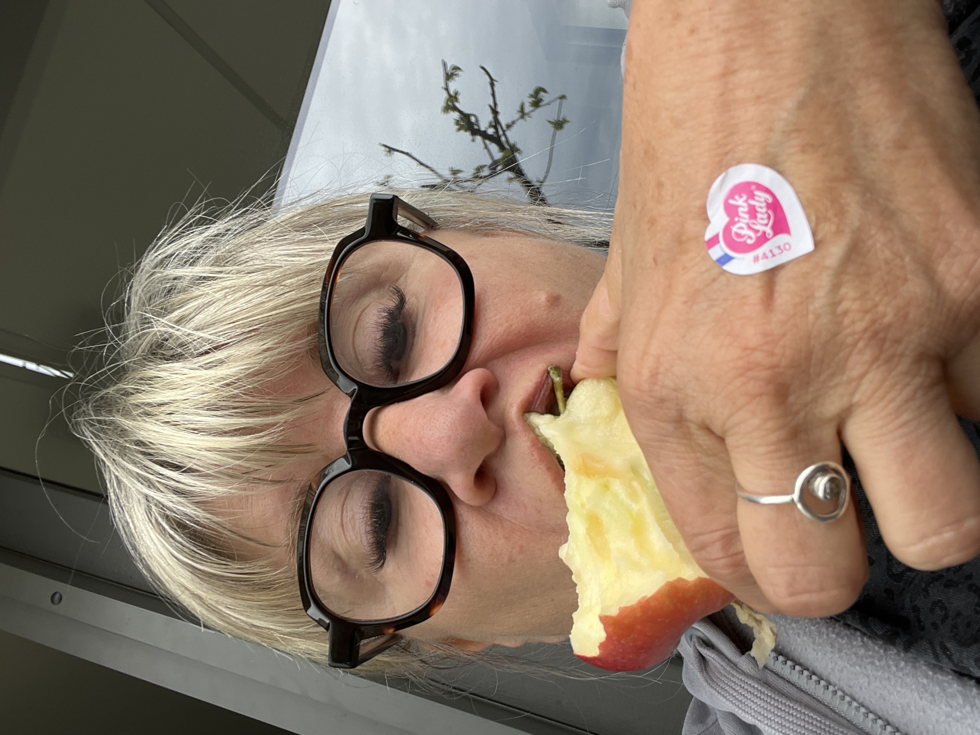 An eye-level shot captures a person, likely a woman with blonde hair and dark-framed glasses, in the act of eating an apple. The image is taken from a slightly upward angle, showing her face in profile as she holds a partially eaten Pink Lady apple to her mouth. Her eyes are visible through the glasses, appearing to look downwards.
A heart-shaped sticker, likely from the apple, is adhered to the back of her left hand, which is holding the apple. The sticker clearly displays the words "Pink Lady" and the number "#4130". A silver ring with a circular design is visible on the middle finger of the hand holding the apple.
The background includes a reflection in what appears to be a window or glass surface, showing bare tree branches with newly sprouted small green leaves against a bright, overcast sky. This suggests an outdoor view, possibly from an indoor setting or a vehicle. Part of her clothing, a grey zipped garment, is visible in the lower right corner. The lighting indicates it is daytime, possibly spring given the budding branches. No specific location cues are visible in the image itself.