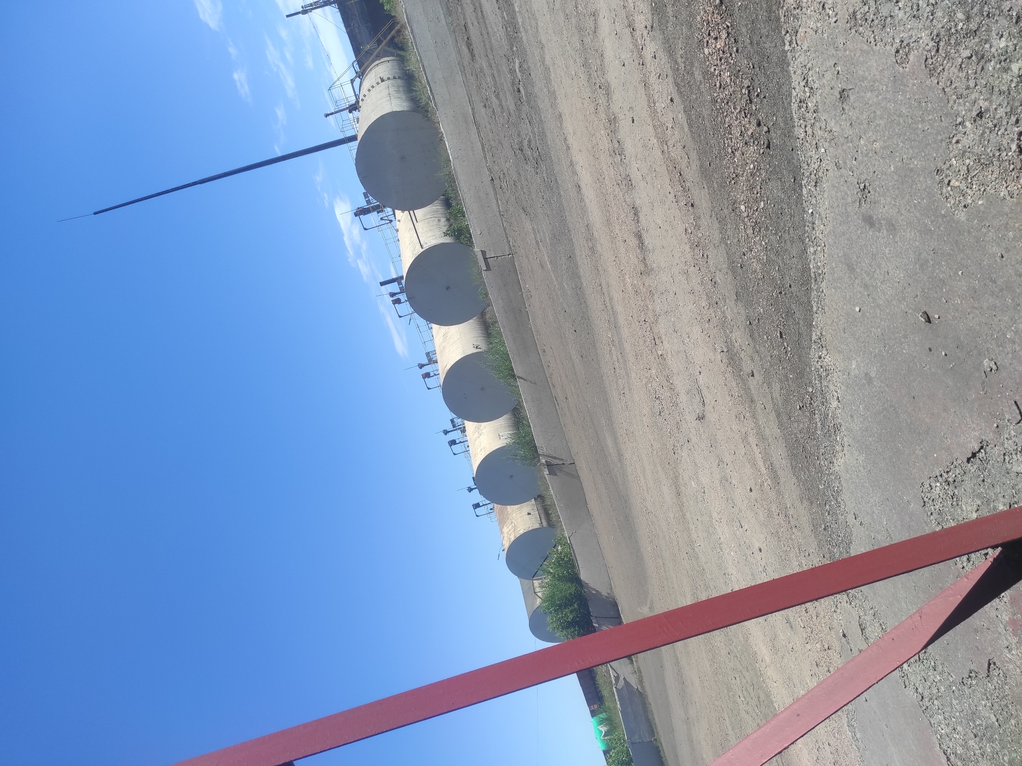 The image shows an industrial area under a clear blue sky with scattered white clouds. In the foreground, a portion of a red metal structure is visible, casting a shadow on a gravelly ground. Further back, a line of large, cylindrical storage tanks, likely for fuel or other industrial materials, are arrayed horizontally. These tanks are supported by a concrete structure and are partially overgrown with green vegetation. A tall, slender dark pole, possibly a lightning rod or communication antenna, extends from behind the tanks towards the sky. The overall scene suggests a remote industrial site within or near the city of Kyzyl, Kazakhstan, characterized by open space, industrial infrastructure, and a bright, sunny day. There are no people or animals visible in the image.