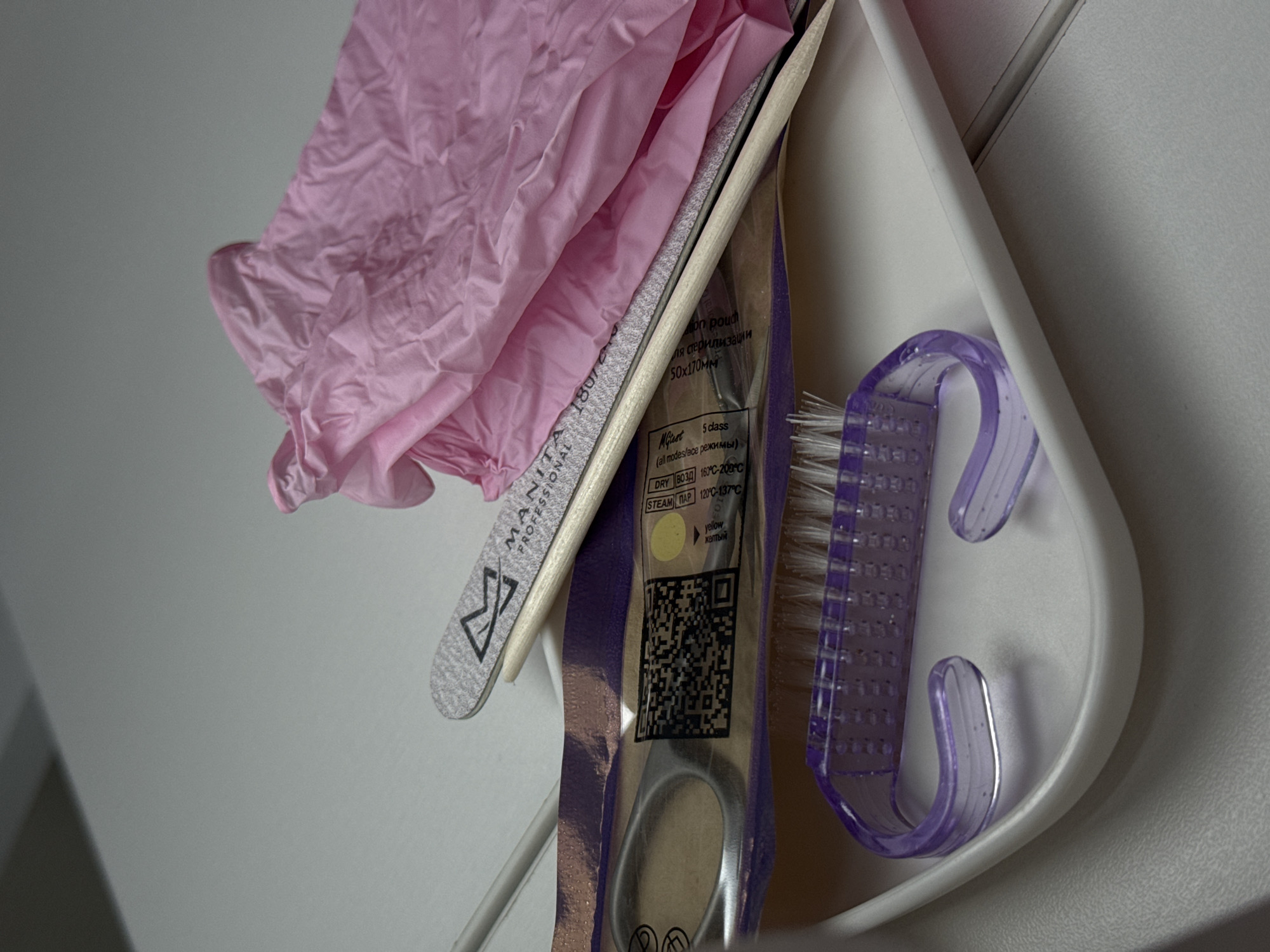 The image shows a collection of manicure tools stored in a white plastic holder. The tools include a pair of scissors in a purple sterile pouch, a nail file labeled "MANITA PROFESSIONAL," a purple manicure brush, and a pair of pink gloves. The tools are likely used for professional nail care, as indicated by the "MANITA PROFESSIONAL" branding and the presence of sterile packaging. The label on the pouch is in Russian, indicating it is likely for use in the city of Chaikovsky, Russia.