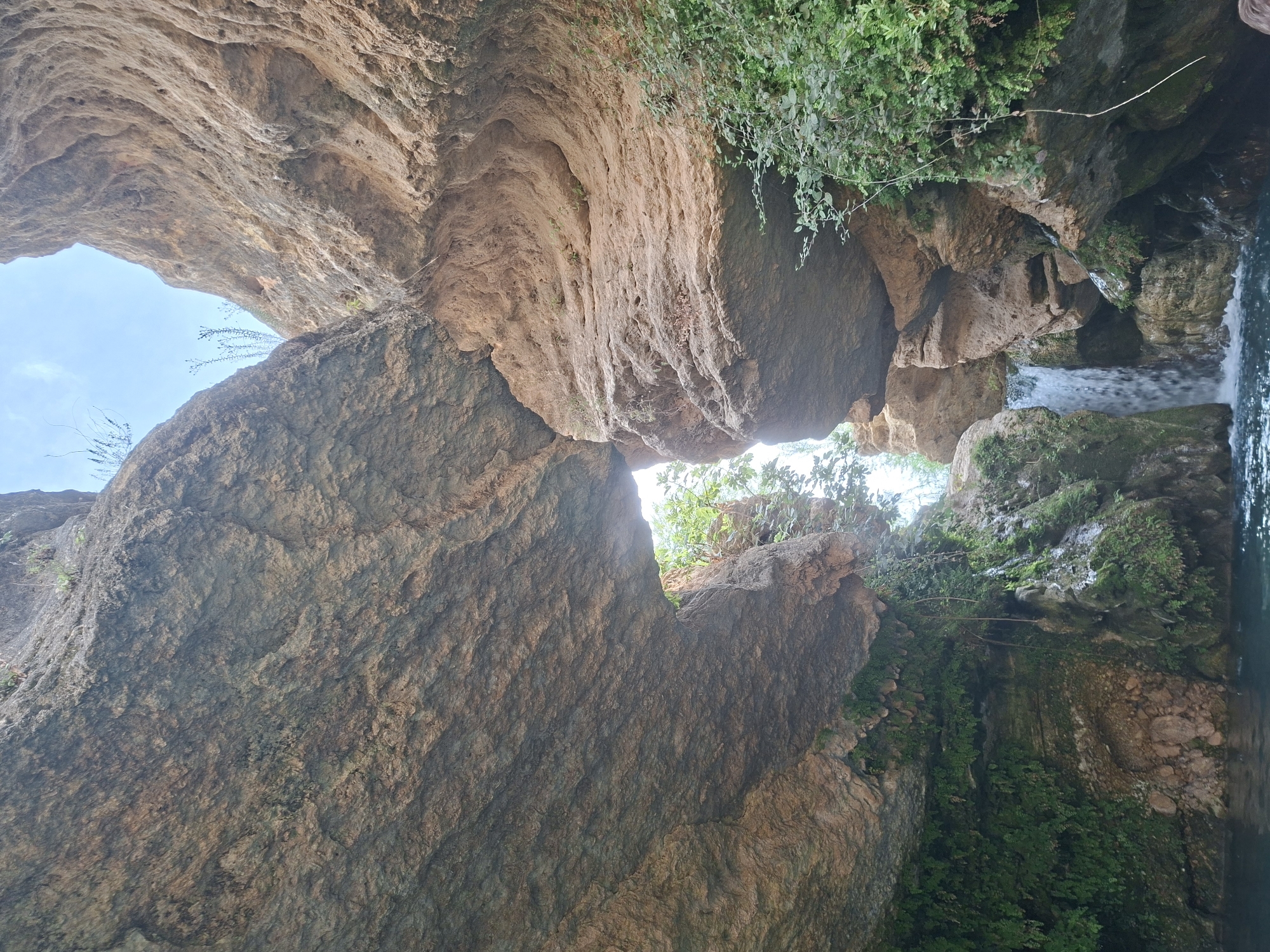 The image depicts a natural scene within a canyon, featuring towering rock formations with rugged textures and earthy tones. Lush green vegetation, including mosses and ferns, clings to the rock faces, particularly on the left side. A small waterfall cascades down the rocks into a pool of dark blue water at the bottom of the frame. Sunlight filters through a gap between the rock overhangs, illuminating the scene. The style of the rock formations and the presence of a waterfall suggest a natural landscape, potentially a ravine or gorge, with elements of water erosion and vegetation growth. The location is identified as Unknown, Spain, indicating a specific but unnamed place within the country. There are no people or identifiable objects that suggest human activity in this particular view, focusing instead on the natural environment.