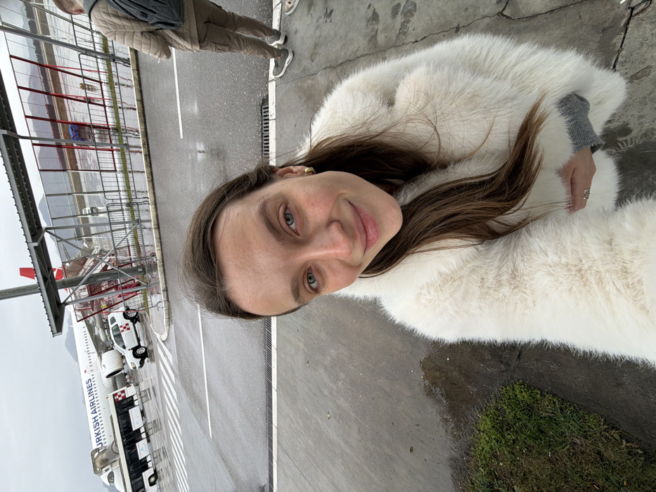 A woman wearing a white fur coat poses for a selfie at an airport. A Turkish Airlines plane and vehicles are visible on the tarmac. The sky is overcast, and the ground appears wet. A second person is partially visible in the background near a glass and metal structure.