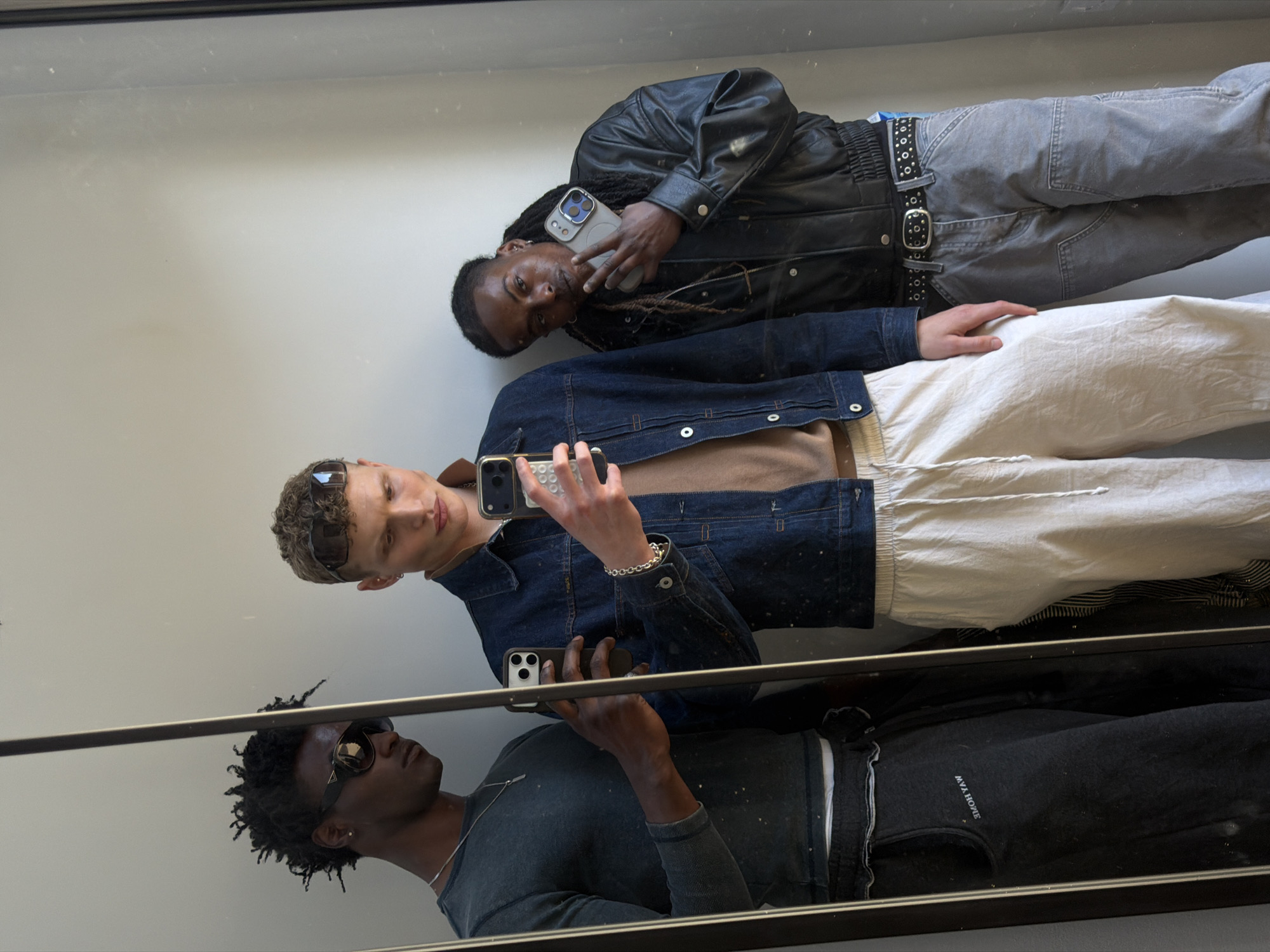 The image displays a mirror selfie featuring three men, likely taken indoors in London, United Kingdom, during daytime given the bright lighting. The reflection shows the men positioned vertically, suggesting they are standing in front of the mirror.
At the top, a man with dark skin and dreadlocks wears a black leather jacket, grey cargo pants, and a black studded belt. He holds a light-colored iPhone to his face and makes a peace sign with his other hand, looking directly into the camera with a relaxed expression.
In the middle, a man with light skin, curly brown hair, and dark sunglasses is captured. He is wearing a dark denim jacket over a light brown top and white, possibly linen, drawstring pants. He holds a dark-colored iPhone, which is actively taking the selfie, and wears a silver chain bracelet. His gaze is directed towards the phone, focused on the act of photography.
At the bottom, a third man with dark skin, short curly hair, and dark sunglasses is partially visible. He wears a dark grey long-sleeved shirt and dark grey sweatpants, on which the text "MVA HOME" is discernible. He is looking away from the camera, giving a more contemplative impression.
The background behind the reflections is a plain, light-colored wall. A dark horizontal bar or frame runs across the lower third of the image, separating the bottom man's reflection from the other two. The overall scene conveys a casual and modern aesthetic.