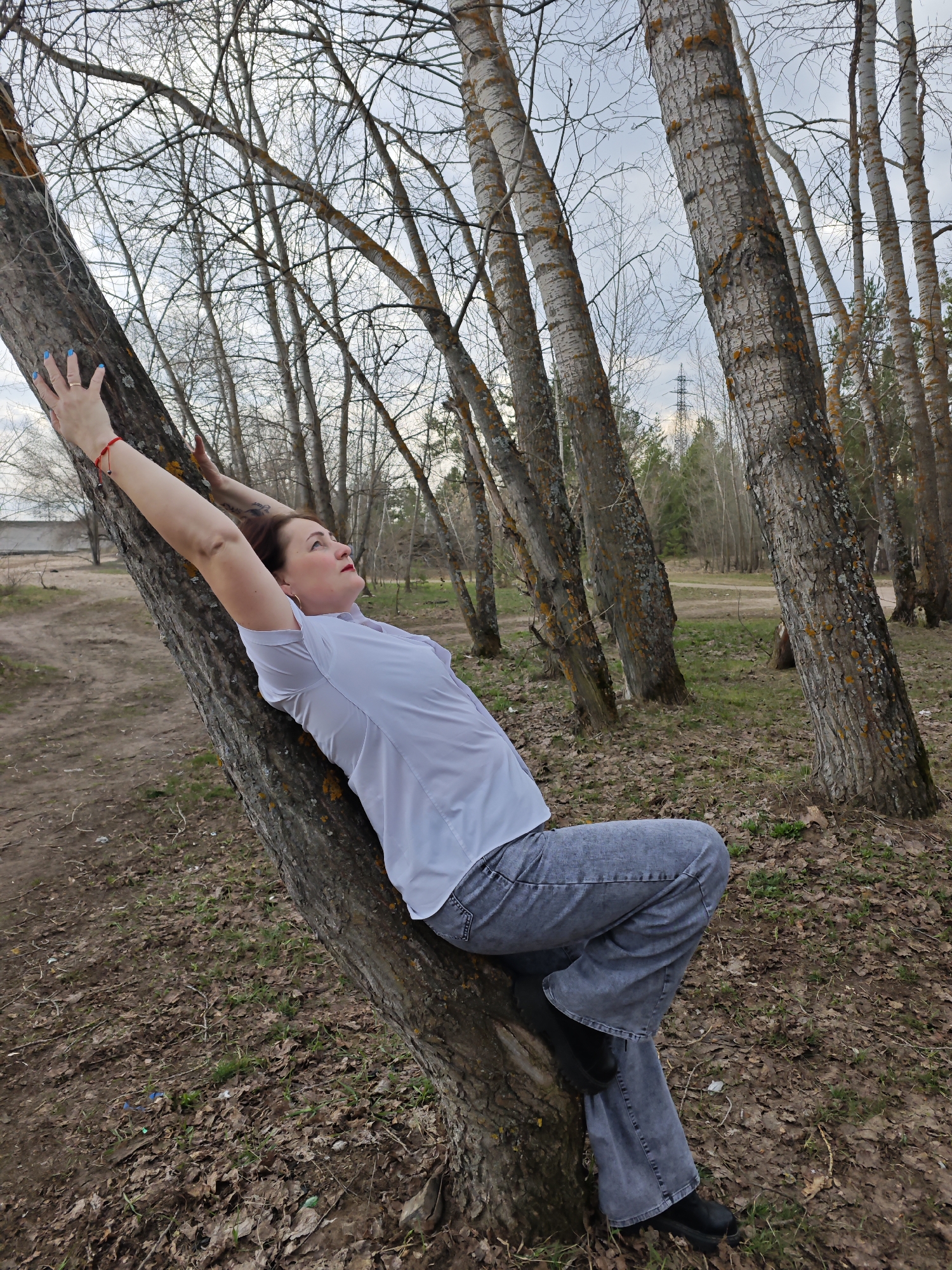 A woman is captured in a wooded setting, leaning against the trunk of a tree and stretching her arms upwards, resting her hands on the bark. She appears to be middle-aged, with dark hair, and is wearing a light-colored, collared short-sleeved shirt, light gray wide-leg jeans, and black shoes. Her left wrist features a red beaded bracelet, and her fingernails are painted blue. She has red lipstick and looks upwards with a serene, contemplative expression. A tattoo is visible on her right forearm.
The scene is set in what appears to be a forest or grove, characterized by numerous trees with bare branches, indicating either late winter or early spring. The ground is covered with dry, fallen leaves and sparse patches of green grass are starting to emerge. Many of the trees have textured, light-colored bark, some showing patches of orange lichen. The sky is overcast, casting a soft, diffused light over the landscape. In the background, a clearing or dirt path is visible, and a power line tower can be faintly seen through the trees, suggesting the area is near human infrastructure. The overall impression is one of calm and natural simplicity.