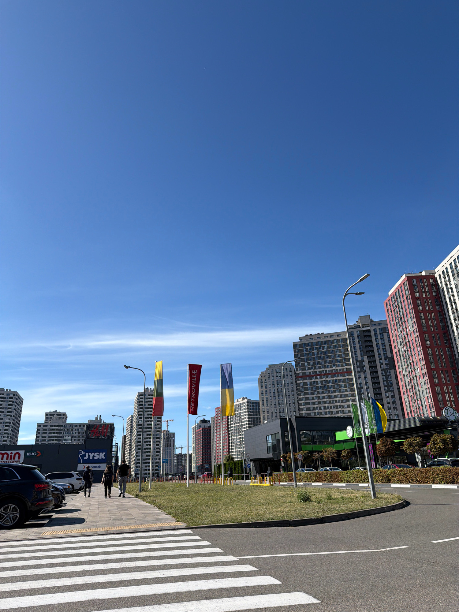 An outdoor, eye-level, wide shot captures a bright, sunny day with a clear blue sky over a commercial area in Kyiv, Ukraine. The foreground features a pedestrian crosswalk with white stripes on asphalt, leading to a sidewalk paved with light-colored tiles. Three individuals are walking away from the camera on the sidewalk towards the right. To the left, a black SUV is parked next to other vehicles. Further into the scene, several modern, multi-story apartment buildings rise into the sky. Some buildings have red facades, while others are gray and white. Flags are visible on poles in front of some buildings, including one red flag with white text that reads "RETROVILLE," and two flags with vertical stripes of yellow, green, and blue, representing Ukraine's national colors. In the background, there are some trees and a construction crane. The environment suggests a developed urban area with residential and commercial elements.
