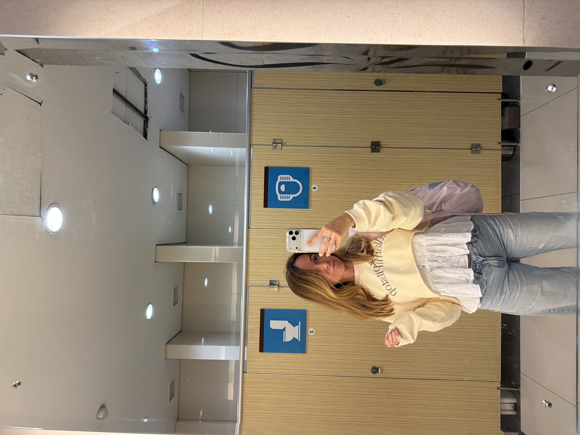 A woman is taking a selfie in a restroom. She is wearing a yellow cropped sweatshirt over a white ruffled shirt and light-wash jeans. She is also carrying a pink backpack. The restroom has wooden stalls with blue signs indicating a toilet and a changing station. The floor is tiled and reflective, showing the ceiling with lights and a section of damaged ceiling tiles. The scene is a typical public restroom environment. The location is not specified in the image, and therefore Kuta, Indonesia cannot be confirmed.