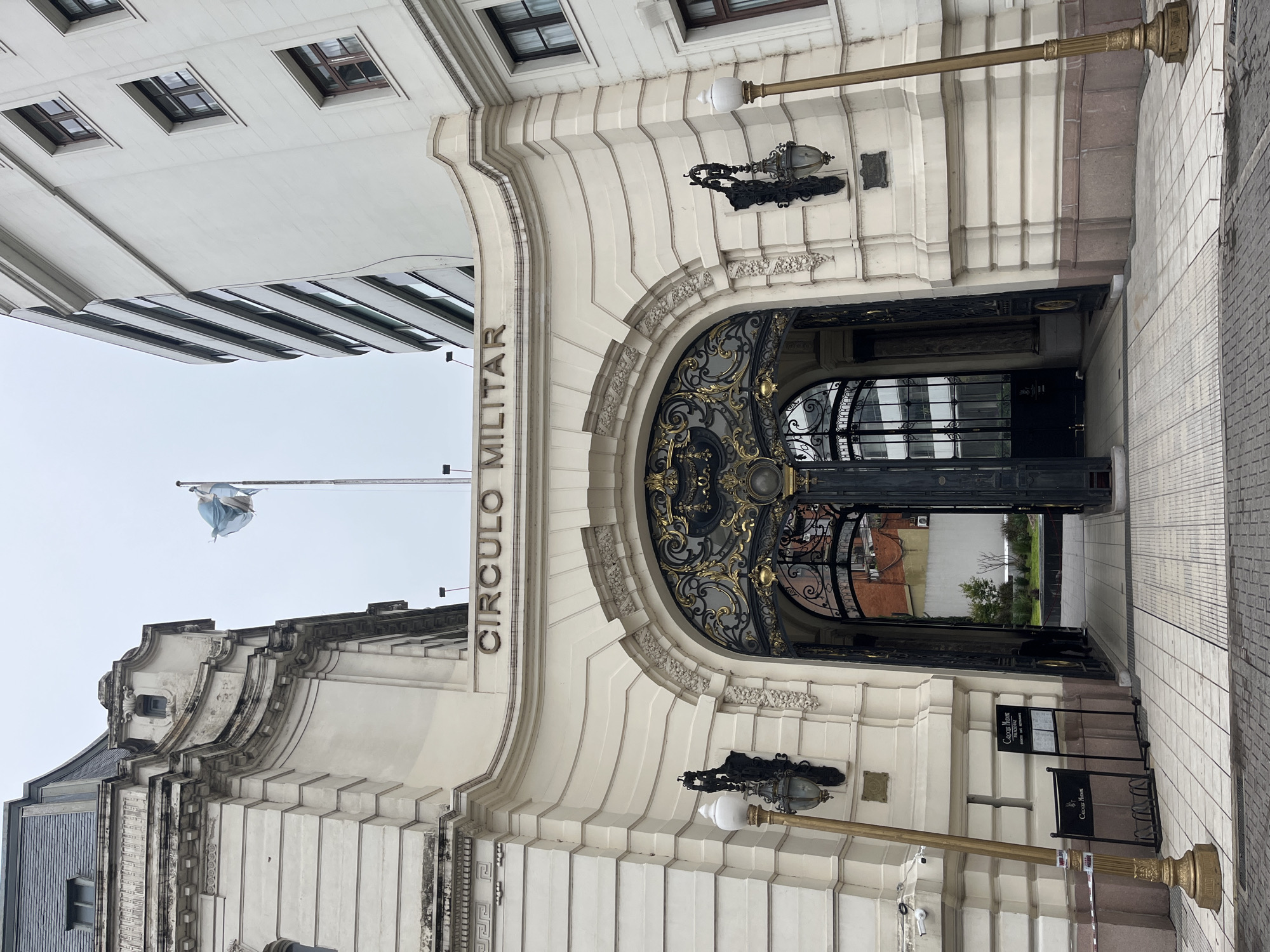 This is a street-level, eye-level, outdoor photograph of the front entrance to the Círculo Militar (Military Circle) building in Buenos Aires, Argentina. The entrance is an arched gateway with an ornate black wrought iron gate, partially open. "CIRCULO MILITAR" is inscribed in block letters above the arch. Two decorative wrought iron lamps flank the archway on the building's exterior. Two black signs for "Cacique House" are visible near the entrance, suggesting a restaurant is located inside. Beyond the gate, there is a view into a courtyard with trees and a building in the background. A flag on a pole is visible to the left of the archway. The building is predominantly white with architectural details and moldings. A tiled sidewalk leads to the entrance. The sky is overcast.