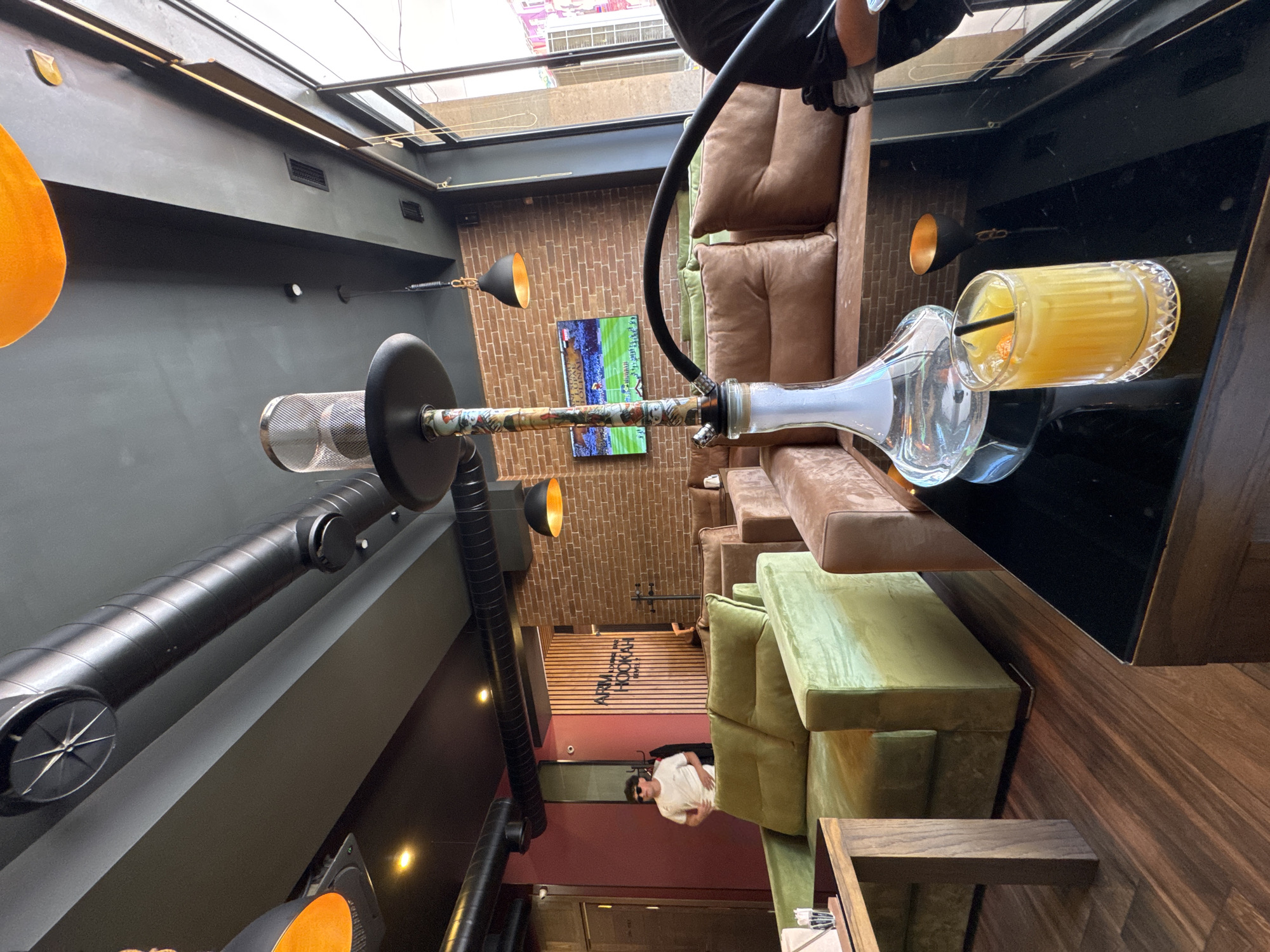 A tilted, high-angle shot reveals the interior of a cafe or lounge, likely in Yerevan, Armenia. The foreground features a table with a hookah and a glass of orange juice with a straw. Seating areas with leather and olive green velvet cushions line the perimeter. Overhead, there is a skylight. A television screen shows a sports game on a brick wall. The ceiling has industrial-style exposed ductwork and light fixtures. A man wearing sunglasses and a white shirt is seen near the entrance, facing forward. Text on a sign reads "ART'L HOOKAH LOUNGE".