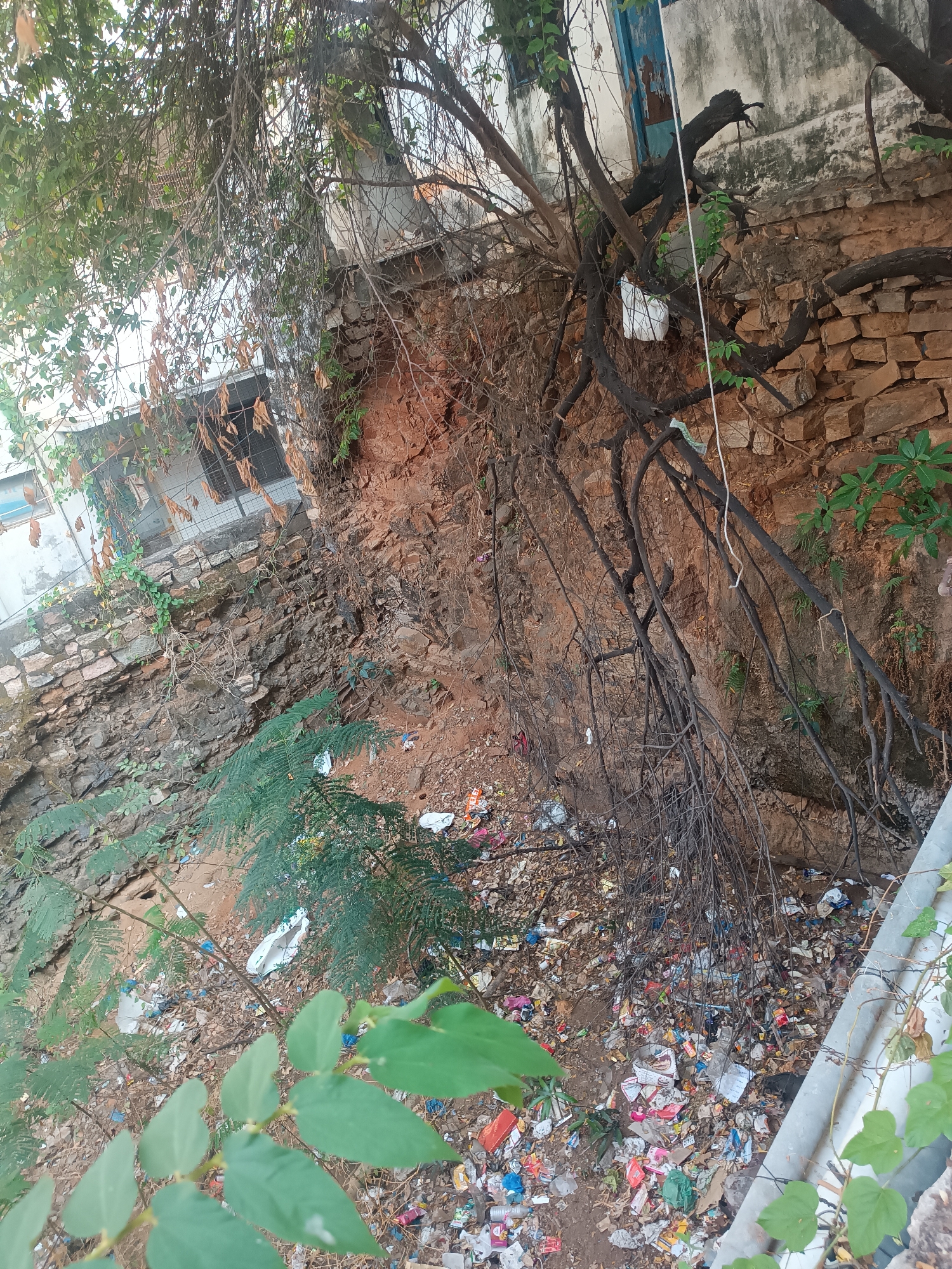 The image depicts a polluted ravine in Puttaparthi, India. A retaining wall of irregular stone blocks supports an eroding bank of reddish-brown soil. A building is visible at the top of the bank. Bare tree branches and some foliage hang over the ravine. The ground is covered with a dense layer of trash, including plastic wrappers, bottles, and other debris. A concrete barrier runs along the edge of the ravine in the foreground. The scene conveys a sense of environmental neglect and urban decay.