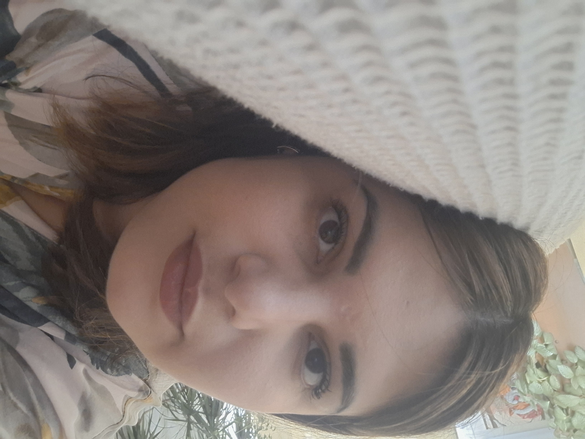 A close-up selfie shows a woman with brown hair and brown eyes. Part of a cream-colored knitted sweater is visible above her head. She is wearing lipstick. A patterned blouse is visible to her left, and green foliage is visible in the background. The location and city of Амзя, Russia, cannot be determined from the image.