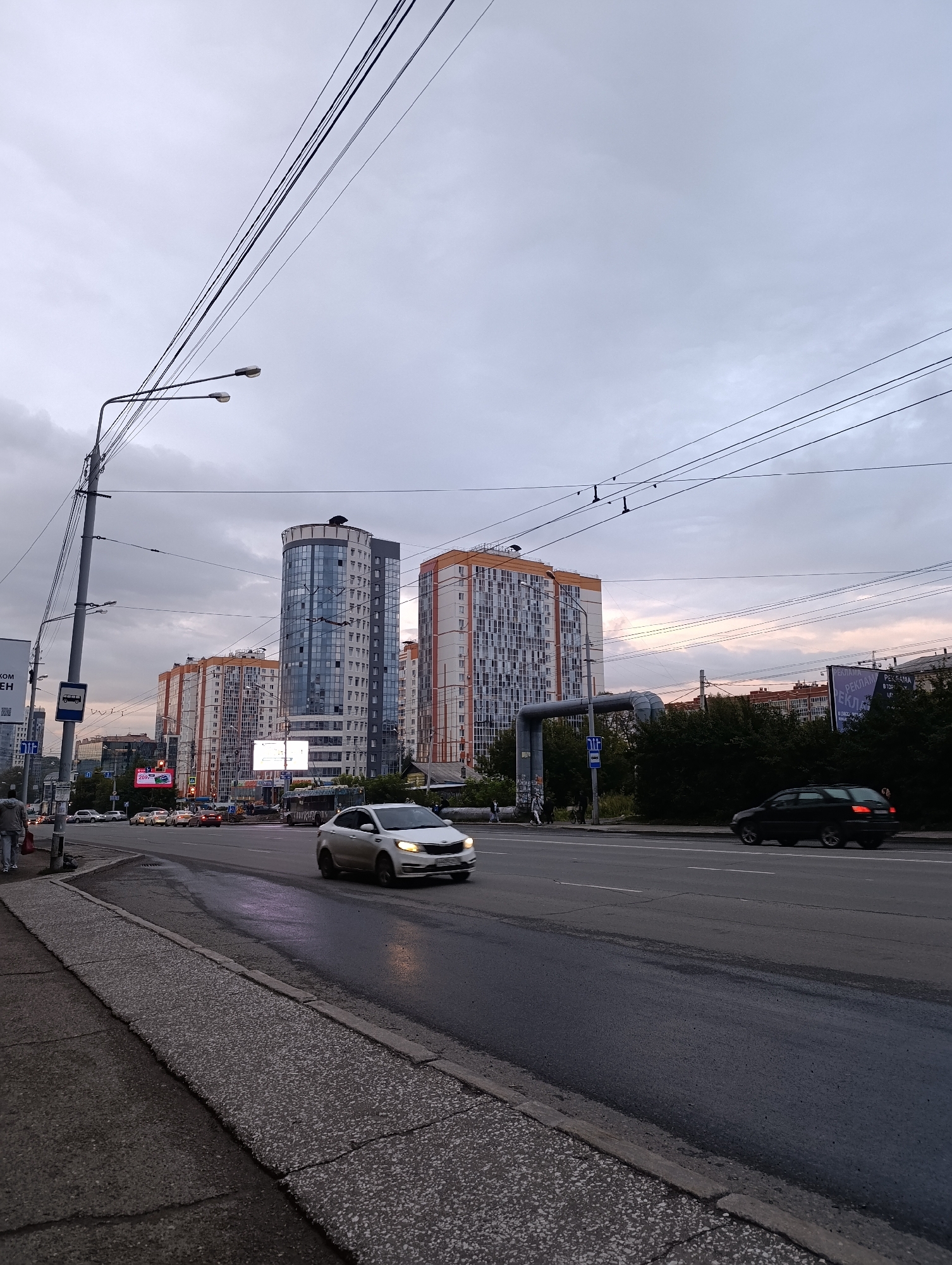 The image captures a street scene in Tomsk, Russia, under an overcast sky with subtle hints of sunset colors. A multi-lane road dominates the foreground, with wet asphalt indicating recent rain. A white sedan is driving in the right lane, its headlights illuminated. A darker sedan is further down the road in the left lane. On the left side of the road, a sidewalk is visible, with a few pedestrians walking. A bus stop sign is also present near the sidewalk.
In the background, a row of modern apartment buildings lines the street. The tallest building, centrally located, is a circular tower with a glass facade. To its right are two taller, rectangular buildings with predominantly orange and white exteriors. Further back, more buildings and trees can be seen. Utility poles and numerous overhead wires crisscross the sky, a common feature in urban environments. Billboards are also visible, one displaying an advertisement. The overall impression is a typical evening urban street scene in a Russian city.