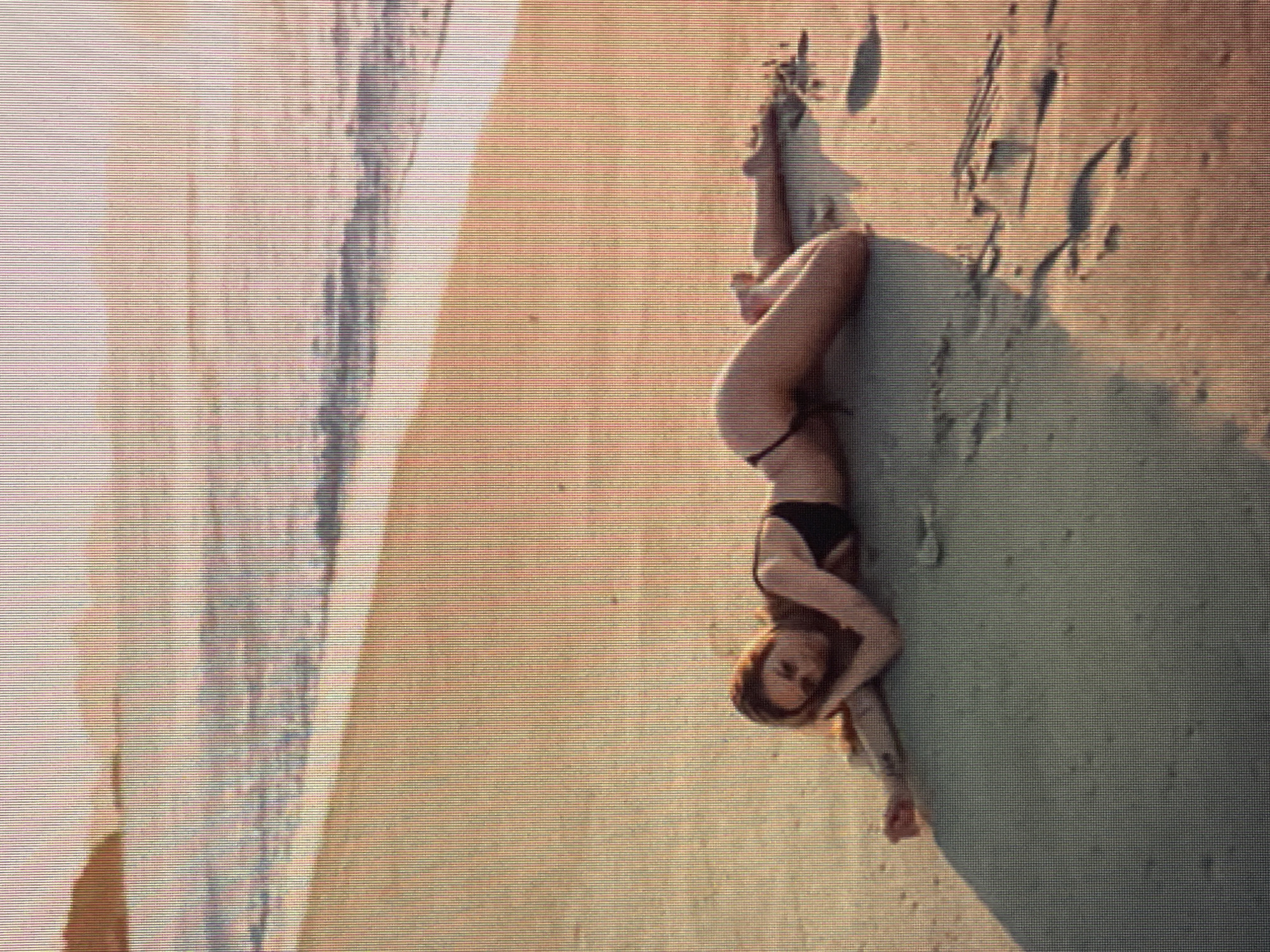 The image shows a person lying on a sandy beach. The person is wearing a black bikini and has long, reddish-brown hair. They are positioned on their side, with their legs bent and their arms resting behind their head. The beach appears to be near the ocean, as evidenced by the water and sand textures. The lighting suggests it could be daytime, possibly late afternoon or early evening, given the warm tones. The sand has some footprints and textures, indicating it is a natural beach environment. The overall mood of the image is relaxed and serene. There is no visible text in the image.