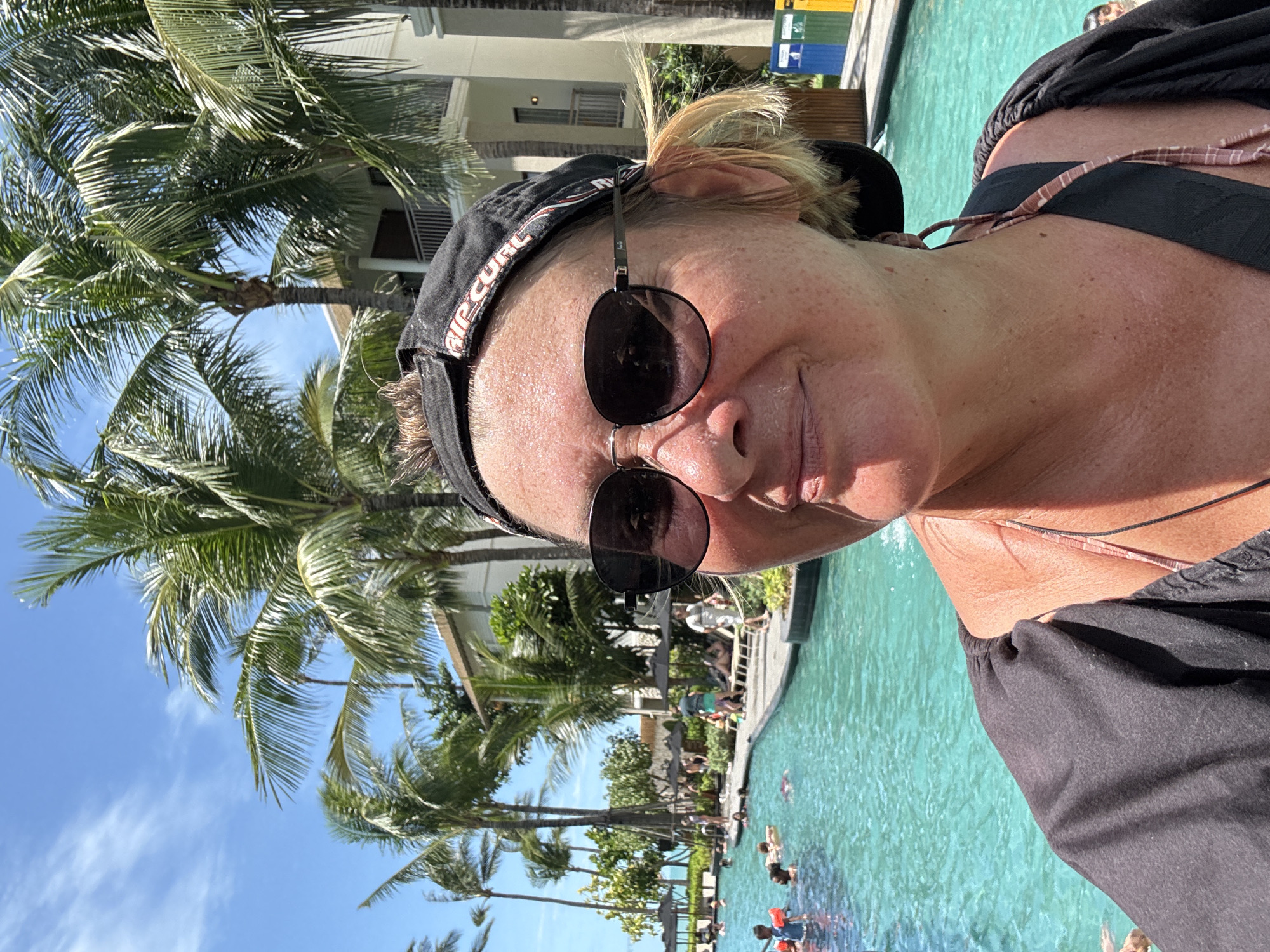 A woman wearing sunglasses and a black cap is taking a selfie next to a swimming pool in Baan Bo Phut, Thailand. Palm trees are visible in the background, along with a building. People are swimming in the pool.
