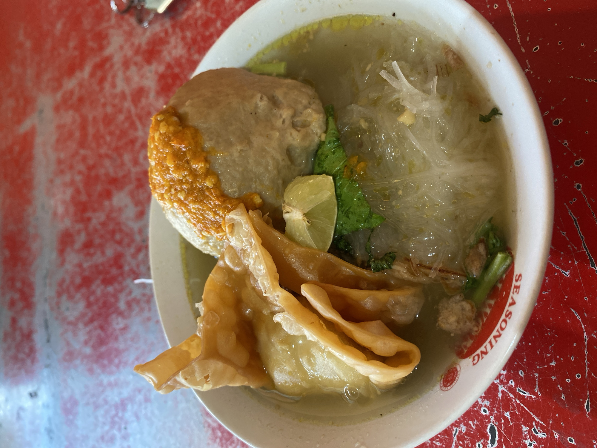 A close-up, overhead shot shows a bowl of Indonesian meatballs, known as "bakso," on a red, textured table. The white bowl has red lettering around the rim that reads "SEASONING" in reverse. Inside the bowl, a large, round meatball is topped with a dollop of orange chili sauce. To the left of the meatball, a golden-brown fried wonton is folded over. The broth contains clear glass noodles, chopped green onions, and small pieces of minced meat. A wedge of lime sits on top of the wonton. The table surface is predominantly red with white and black speckles, suggesting a worn or distressed finish. There are no people visible in the image.