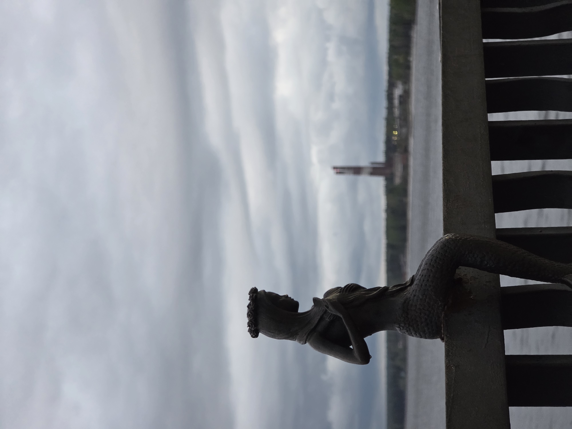 The image shows a mermaid statue located on a bridge in the city of Chaikovsky, Russia. The statue is made of metal. In the background, there is a body of water and an industrial complex, possibly a factory with two smokestacks. The sky is overcast and cloudy.