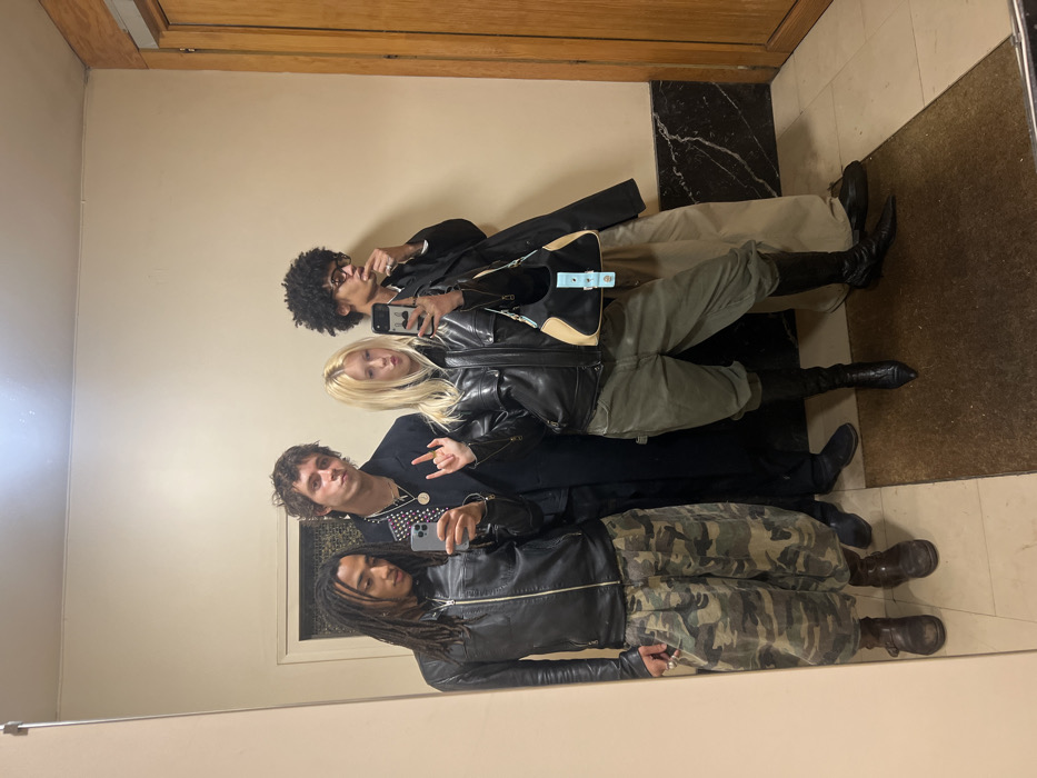 Four people are reflected in a mirror. The first person has dark curly hair, glasses, and wears a dark jacket, beige pants, and black boots. They hold a phone. The second person has blonde hair and wears a leather jacket. The third person wears a patterned shirt, a dark jacket, and makes a hand gesture. They also hold a phone. The fourth person has dreadlocks and wears a leather jacket and camouflage pants. The background includes a wooden door, a beige wall, and a floor with a dark rug.