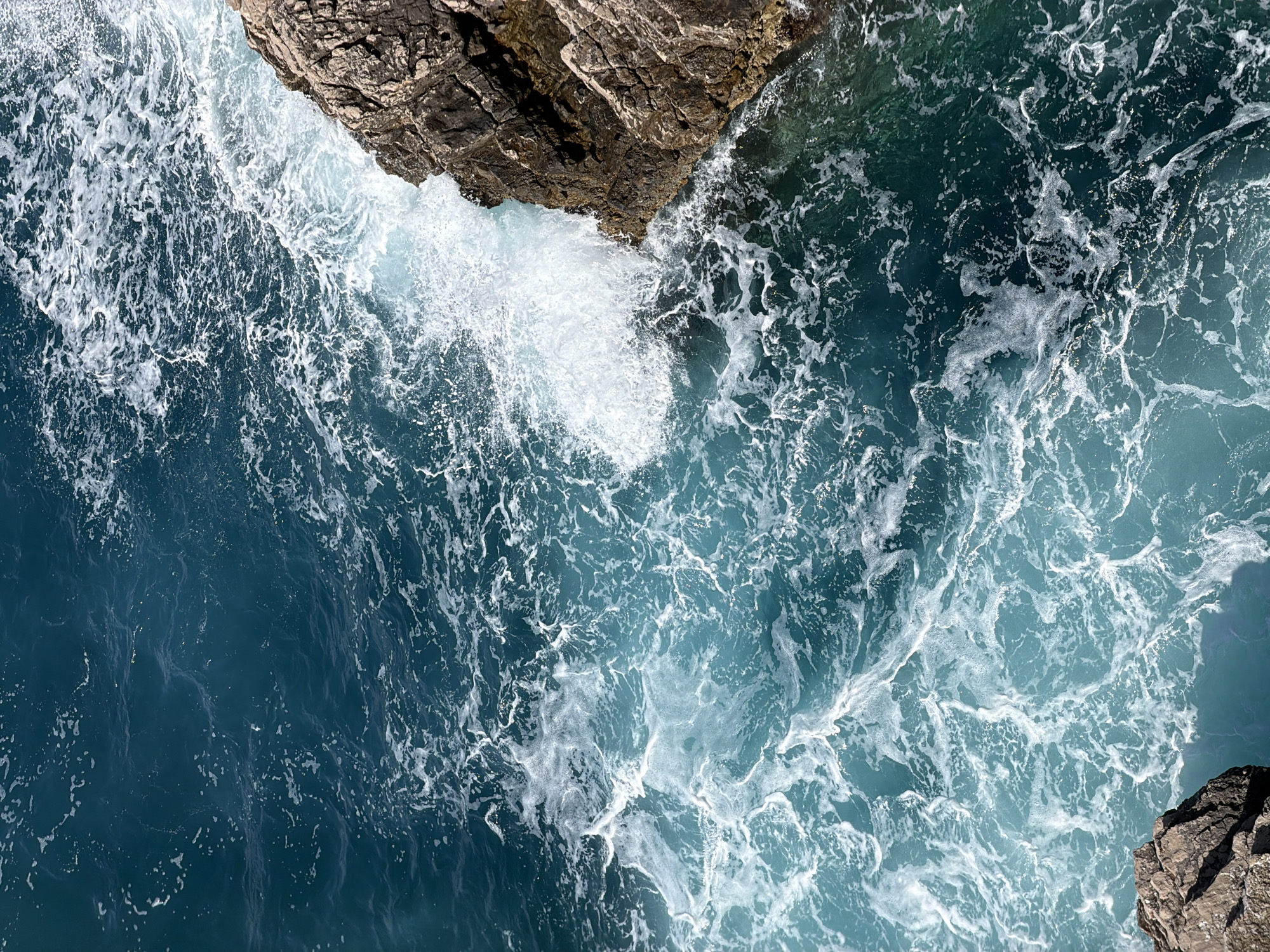 This is an overhead, high-angle view of the sea crashing against a rocky coastline, captured in Mirište, Montenegro. The image is dominated by the turbulent blue-green ocean water and the dark, rugged rock formations.
Jagged, dark brown and grey rocks are visible in the upper left and lower right sections of the frame, showing textured surfaces and receiving direct sunlight, casting some shadows. The main activity depicted is the vigorous movement of waves and sea foam. A significant burst of white foam and spray is prominent where the water forcefully collides with the rocks, particularly in the central-left area.
The water ranges in color from deep, clear blues in calmer areas to vibrant turquoise and teal where it churns and mixes with light. Extensive patterns of white sea foam and bubbles are spread across the water's surface, indicating strong currents and wave action. These foamy trails create intricate, swirling designs that contrast with the deeper hues of the water.
The scene conveys a sense of dynamic natural power, with the ocean's energy continuously shaping the rocky shore. The bright illumination suggests it is daytime, likely under sunny conditions. There is no visible text in the image.