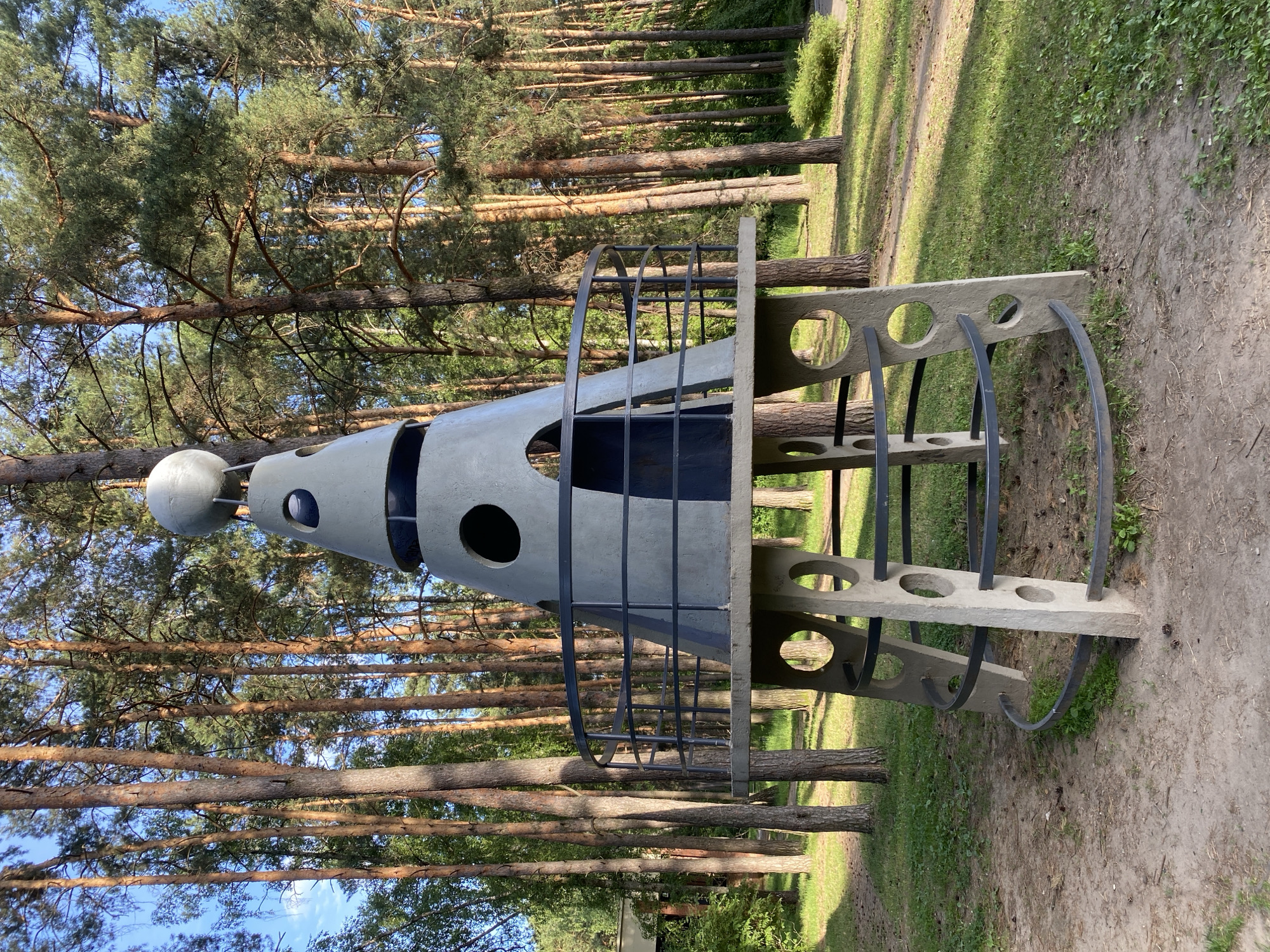 A concrete sculpture resembling a rocket ship stands in a clearing surrounded by a dense forest of tall pine trees. The sculpture is gray and has a conical top adorned with a silver sphere. It features large, circular openings on its conical section and a circular platform with a black metal railing. The base of the sculpture is supported by four concrete pillars, each with several circular cutouts, and is encircled by a black metal railing. The ground around the sculpture is a mix of dirt, grass, and fallen pine needles. The sky above is blue with a few scattered clouds. The overall impression is one of a quirky, slightly retro art installation set within a natural, wooded environment. This sculpture is known to be located in the city of Protvino, Russia, in the Moscow Oblast.