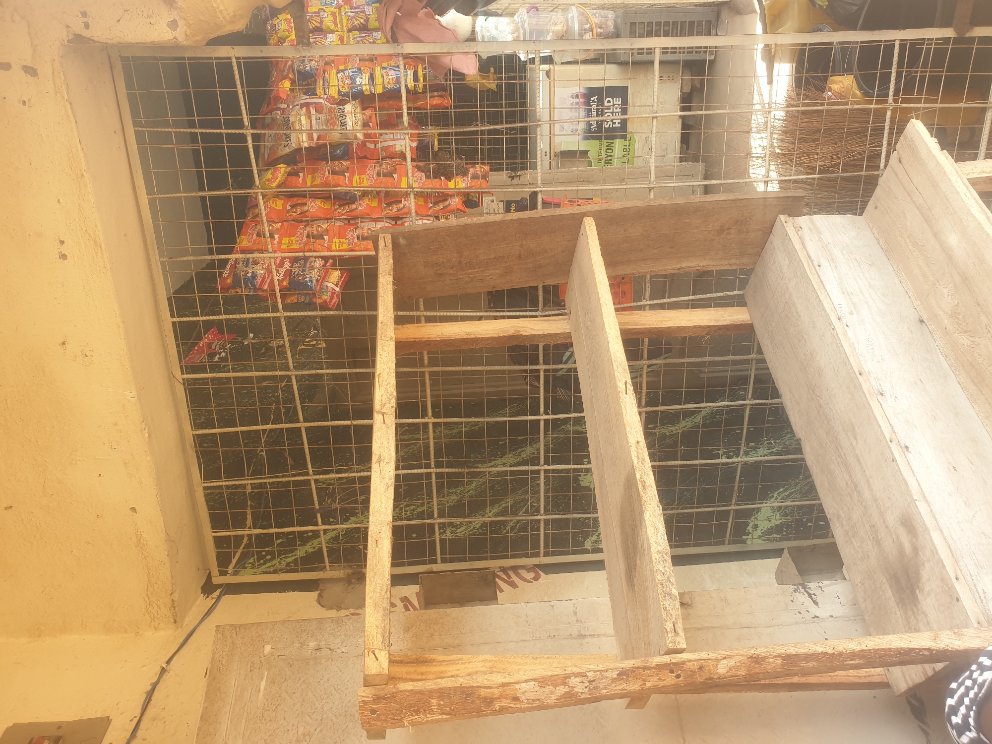 The image depicts an outdoor scene in Jauro Yinu, Nigeria, possibly a small shop or market stall. In the foreground, a rustic, unfinished wooden shelf unit is positioned diagonally. The shelves are wide and made of planks. Behind the shelving unit, a barred window or opening reveals shelves stocked with various packaged goods, primarily snacks like chips or biscuits, in brightly colored wrappers. Some of these packages display prominent brand names such as Kellogg's. A sign on a white background with green lettering, that partially reads "Admiral'A SHORT", "INTERNE ERYON", and "SOLD HERE", is visible through the bars. To the right of the window, a few plastic containers, some filled with goods, are stacked. A bundle of dry grass, possibly a broom, leans against the shelving unit. The overall environment appears to be an outdoor, possibly urban setting, with evidence of construction or makeshift structures suggested by the unfinished wooden shelving. The lighting suggests daytime.