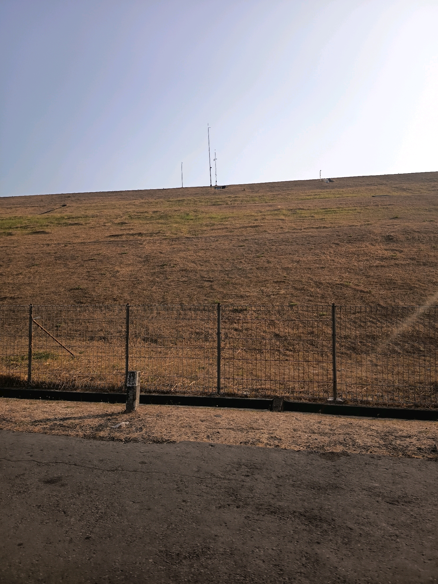 The image shows a wide, grassy embankment, likely part of a dam or levee, under a clear sky. The grass appears dry and brown, suggesting a dry season. A wire fence runs horizontally across the middle ground, separating the embankment from a paved surface in the foreground. Several thin, vertical poles, some with what appear to be antennas or weather instruments, are visible on the crest of the embankment. A single concrete post with some faded markings stands near the fence in the left foreground, casting a distinct shadow. The overall scene is one of a utilitarian outdoor environment, possibly on the outskirts of Kalangbancar, Indonesia.